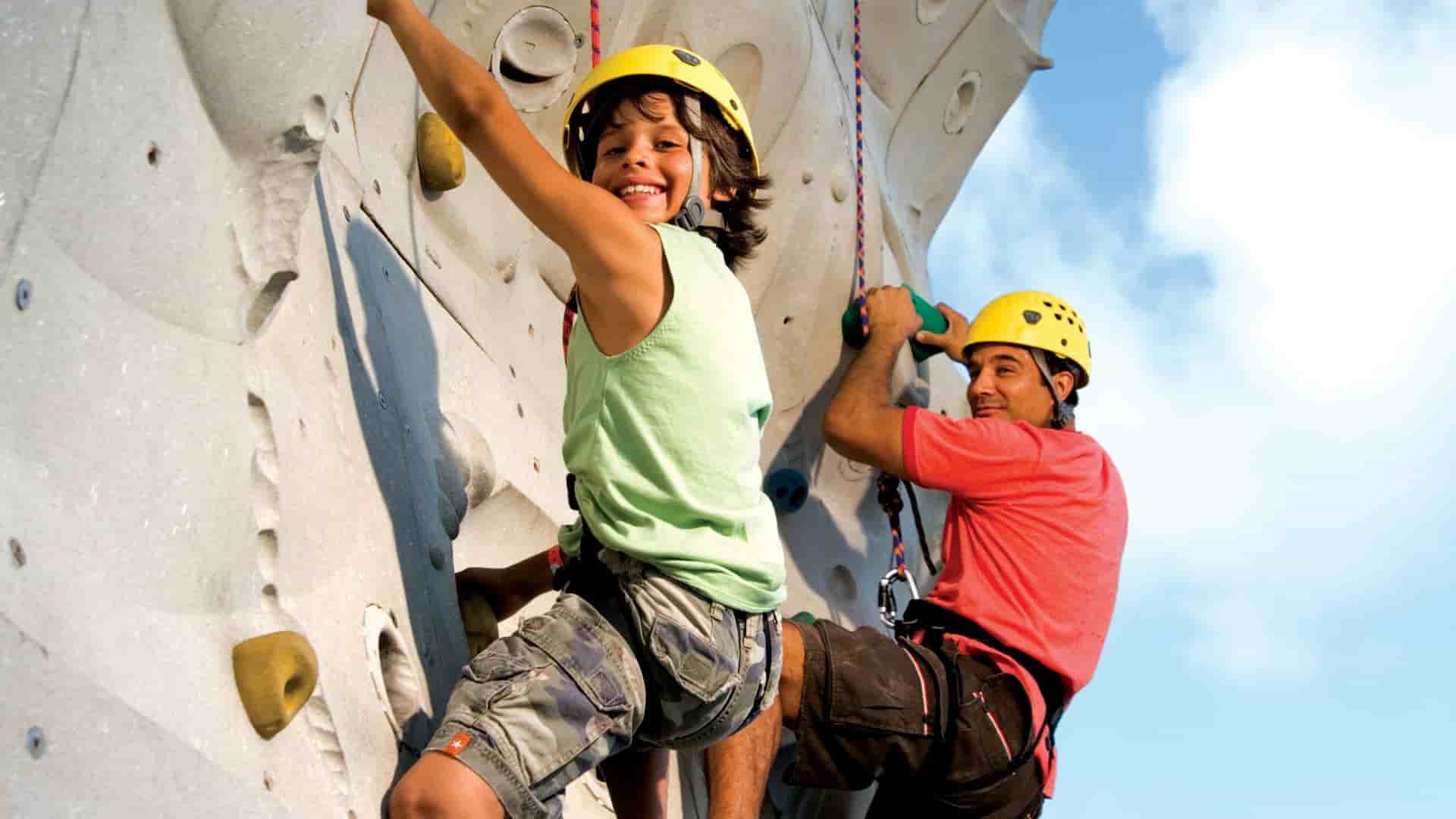 A boy in a yellow helmet and green shirt and a man in a red shirt and yellow helmet are rock climbing outdoors on the Royal Caribbean International Voyager of the Seas ship.