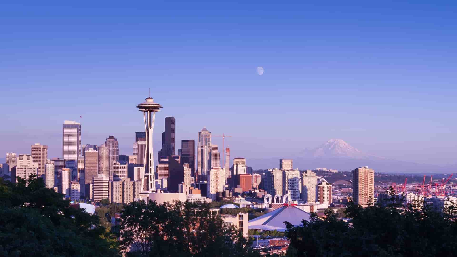 Seattle skyline featuring tall buildings with Mount Rainier in the background and a bright moon in the night sky.