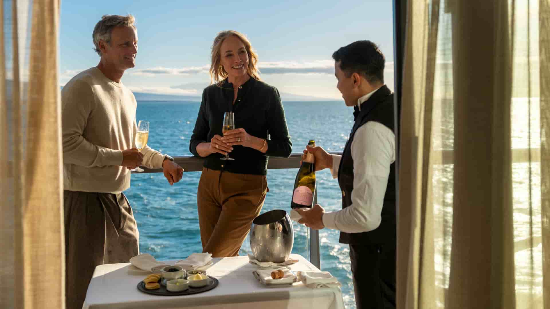 The image shows a butler in a formal uniform pouring champagne for a couple on the private veranda of a Silversea suite. The balcony overlooks a calm, blue ocean under a clear sky, with land visible in the distance. A small table is set with appetizers, a chilled ice bucket, and white linens, illustrating the personalized service and ultra-luxury atmosphere characteristic of Silversea Cruises.
