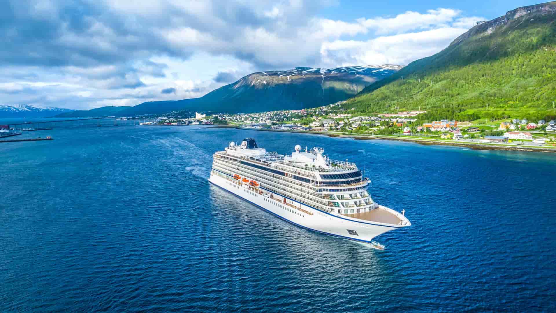 An aerial view of a Viking Ocean cruise ship sailing past the scenic coastal town of Tromsø, Norway, a destination on a Trans-Atlantic itinerary.