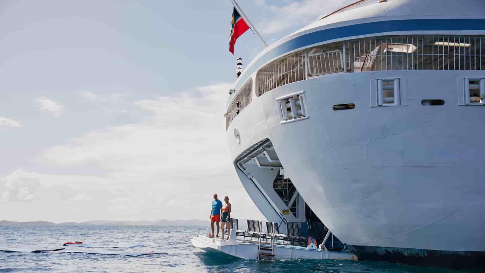A man and woman stand on a cruise ship's retractable watersports marina platform, looking out over the open ocean where floating mats drift nearby.