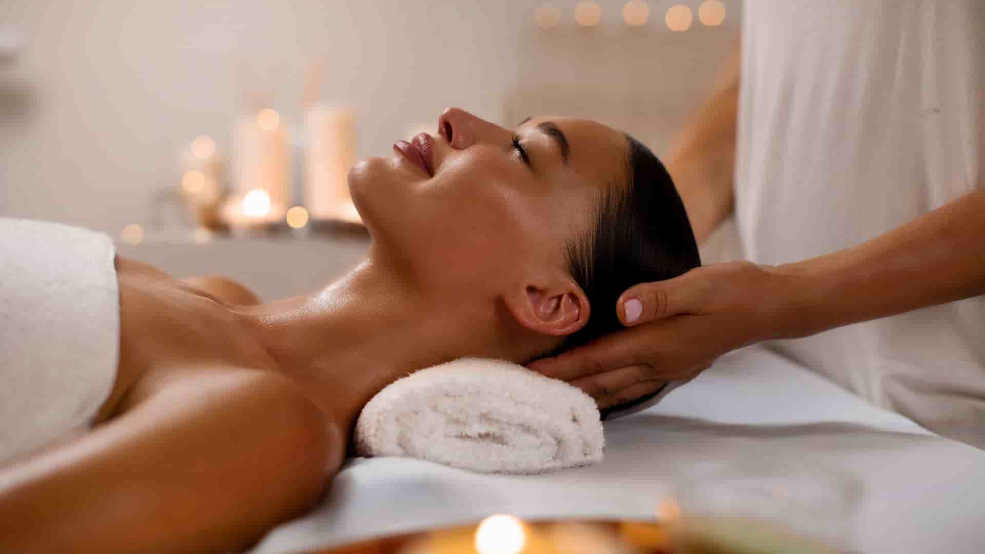 A serene scene of a woman relaxing during a head massage in a spa. She is lying on a massage table with her eyes closed, her hair pulled back, and her shoulders draped in a white towel. A professional massage therapist is gently cradling her head with both hands. The background is softly lit with several candles, creating a warm and tranquil atmosphere. The image captures the relaxing and luxurious experience often found on cruise ships and in spas around the world.