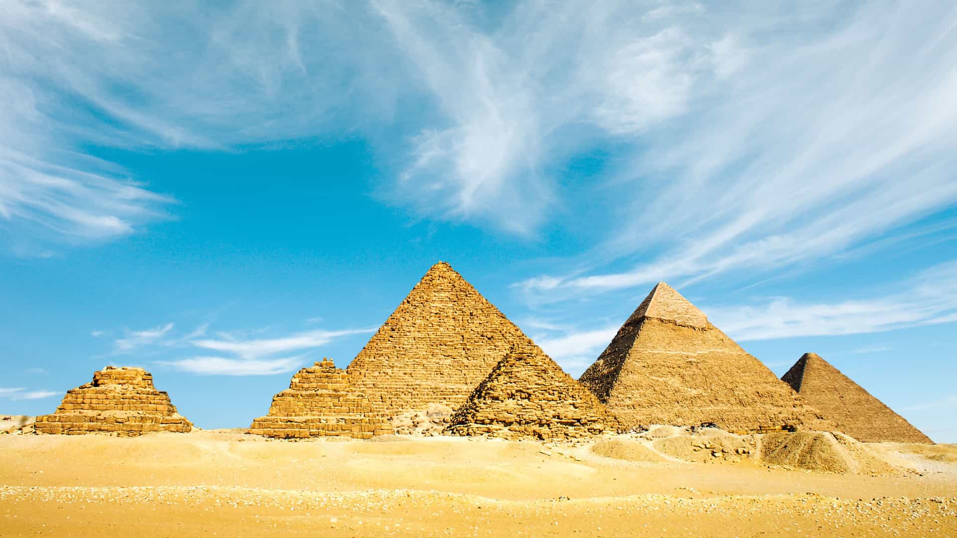 The iconic Pyramids of Giza, Egypt, standing majestically under a bright blue sky with wispy clouds. The massive ancient structures, built from light brown stone, are surrounded by the golden sands of the desert. The image captures the grandeur and historical significance of this famous African landmark.