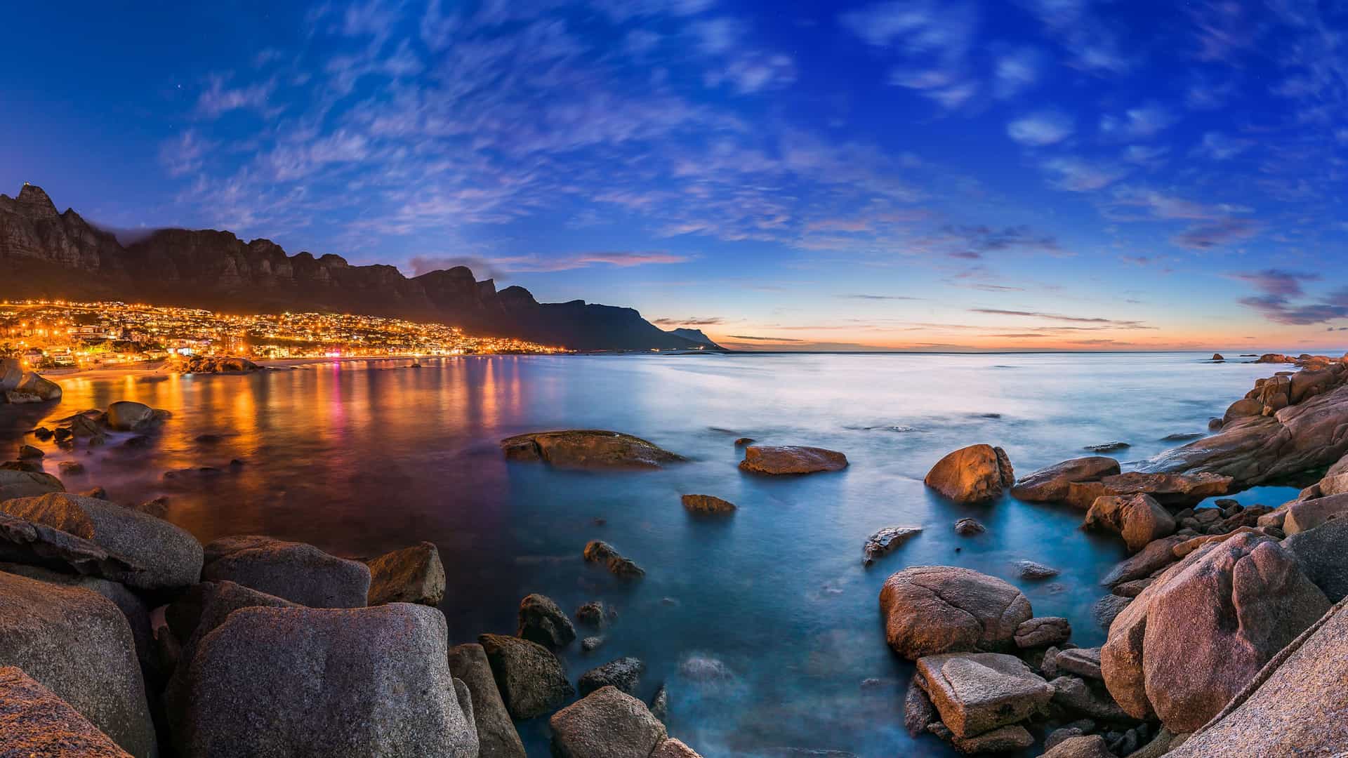 A stunning panoramic view of Cape Town's coastline at dusk, featuring the iconic Table Mountain and the Twelve Apostles mountain range. The city lights glow brightly along the shore, reflecting on the calm ocean. In the foreground, large, dark rocks line the water's edge. The sky is a dramatic blend of deep blue, orange, and purple hues, capturing the beautiful transition from day to night in this famous South African city.