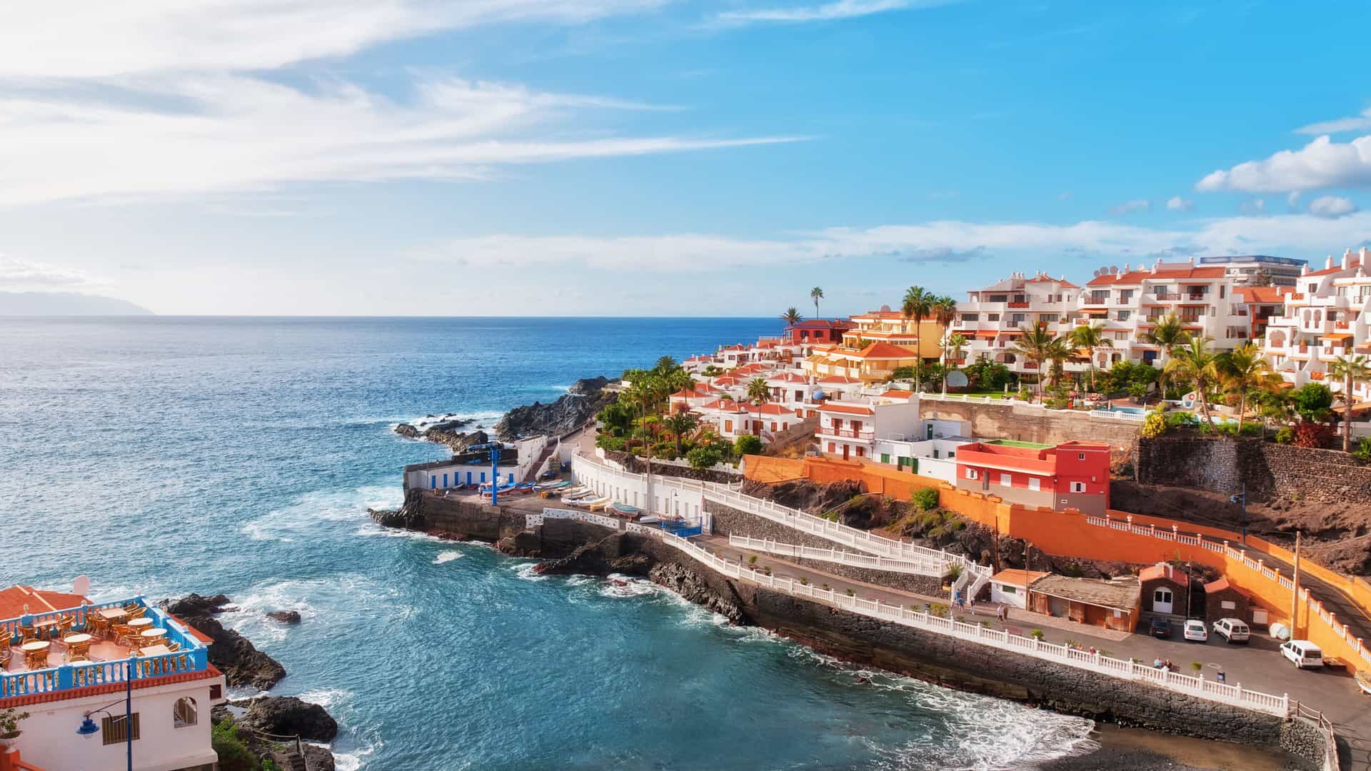 A beautiful view of the coastline of Puerto Santiago in Tenerife, Canary Islands. Colorful buildings with white and red roofs are built into a rocky hillside overlooking the Atlantic Ocean. A paved walkway lines the coast, and a small beach is visible at the bottom right. The water is a mix of deep blue and white foam from the waves crashing against the rocks. The sky is a bright blue with scattered clouds, capturing the scenic beauty of this African island.