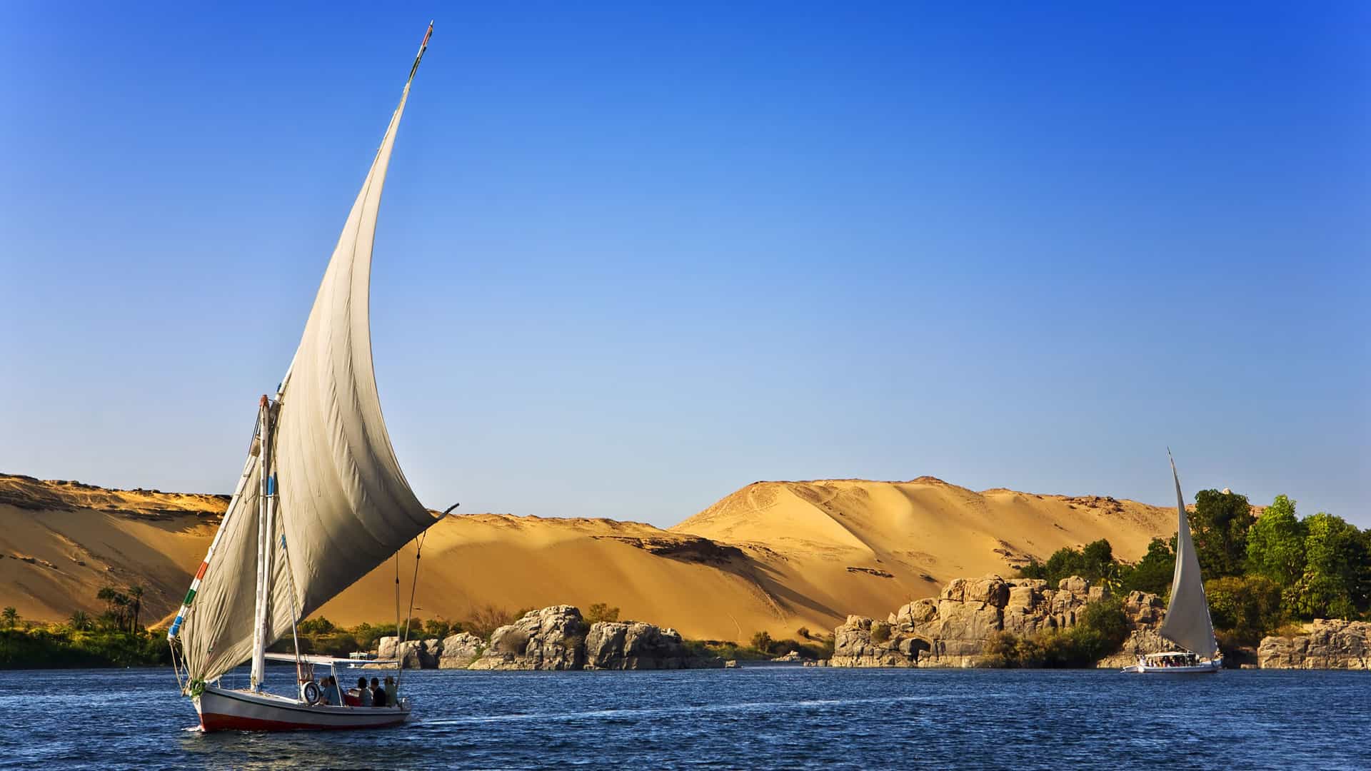 A traditional Egyptian felucca sailboat with a large, white sail glides across the deep blue waters of the Nile River near Aswan. The banks of the river are lined with golden desert dunes on one side and rocky outcroppings and green foliage on the other. Another smaller sailboat is visible in the distance. The sky is a clear, vibrant blue, and the scene captures a peaceful day on the famous Nile River in Africa.