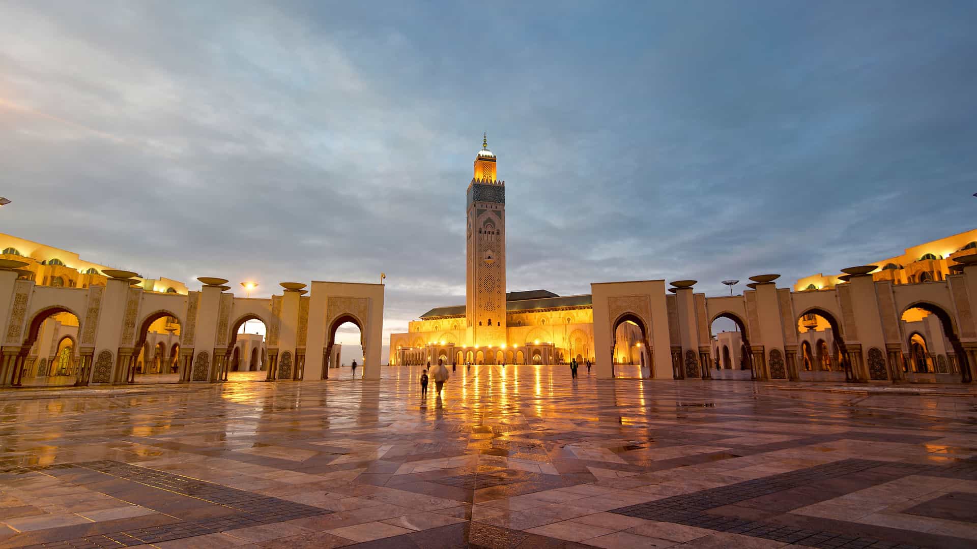The majestic Hassan II Mosque in Casablanca, Morocco, captured at dusk. The grand structure is illuminated with warm yellow lights, highlighting its intricate architecture and soaring minaret. The large, reflective courtyard in the foreground is wet from rain, mirroring the building's lights and the dramatic, cloudy sky. The scene captures the beauty and scale of this iconic African landmark at night.