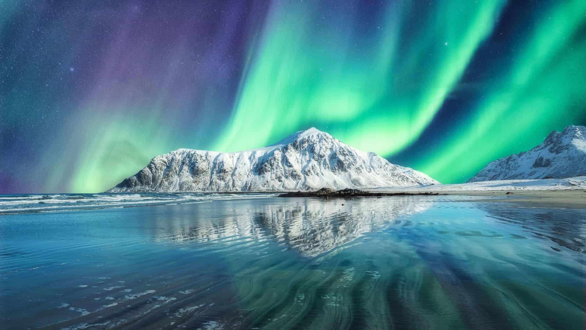 The spectacular Northern Lights, or Aurora Borealis, paint the night sky with vibrant green and purple hues over a snow-covered mountain. The mountain and the celestial display are perfectly reflected in the calm, shallow water of a beach. The foreground shows rippled sand and calm water. This breathtaking image captures the magic of the Arctic Circle region in Norway's Lofoten Islands.