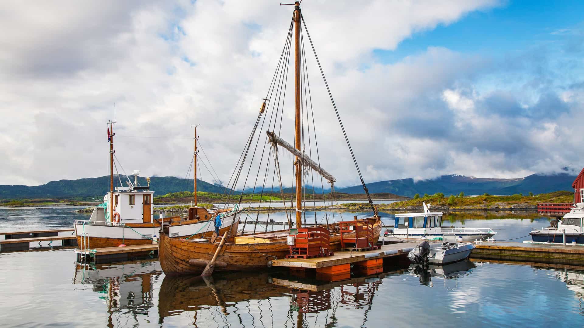 A serene harbor scene on Haholmen Island, Norway, in the Arctic Circle region. Several boats are moored to a wooden dock, including a large, traditional wooden sailing vessel with a tall mast. The water is calm and dark, creating a clear reflection of the boats and the cloudy sky. In the background, rolling green hills rise from the water's edge under a blue sky filled with puffy white clouds, showcasing the peaceful atmosphere of a Norwegian fishing village.