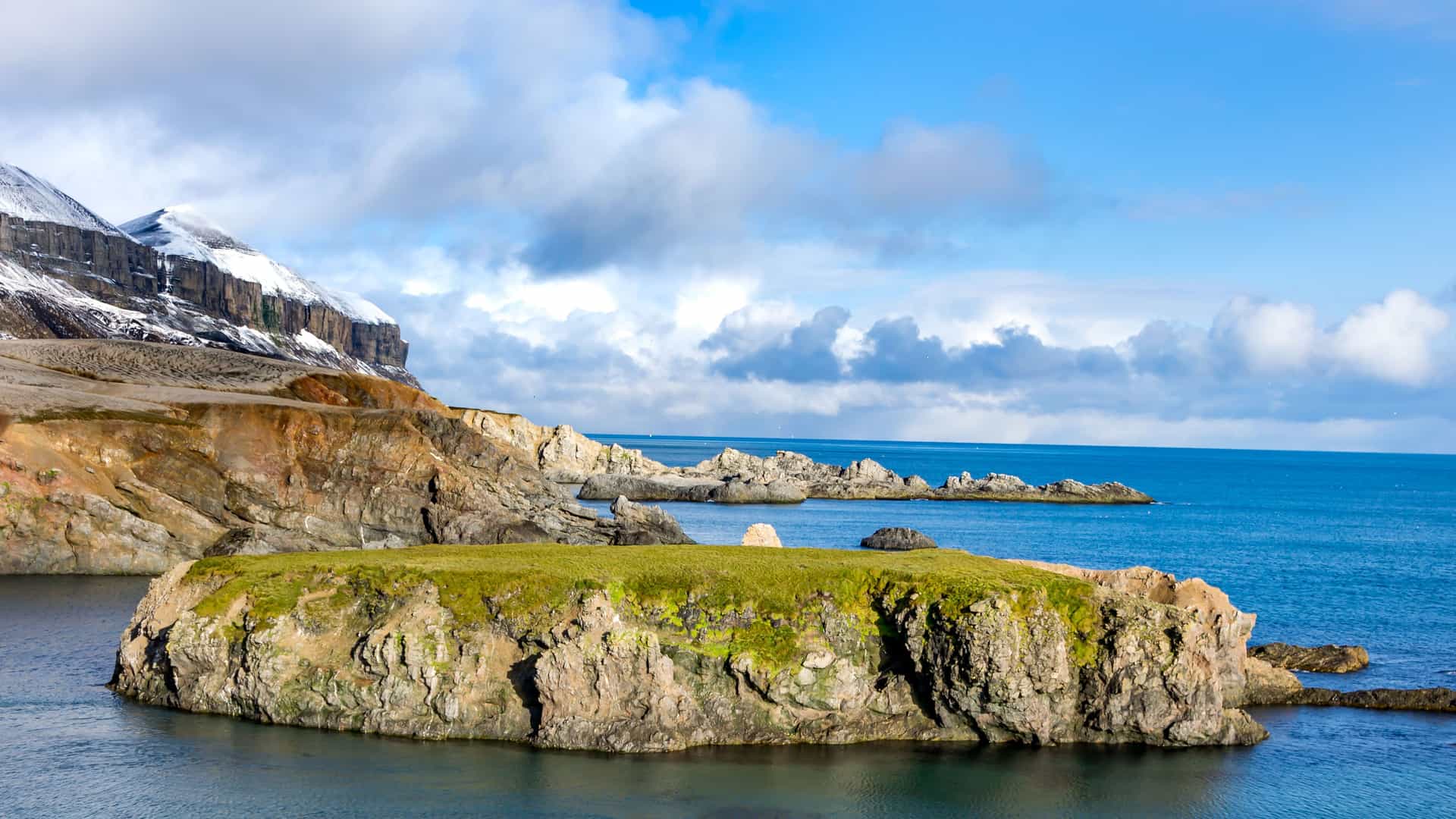 The rugged coastline of Bear Island, Norway, in the Arctic Circle. The image shows a small, rocky island covered in green moss and grass, with a large cliff face rising from the water's edge in the background. The cliff is a mix of brown rock and snow-covered peaks. The sea is a deep blue, and the sky is a bright blue with scattered white clouds, depicting the stark and beautiful arctic landscape.