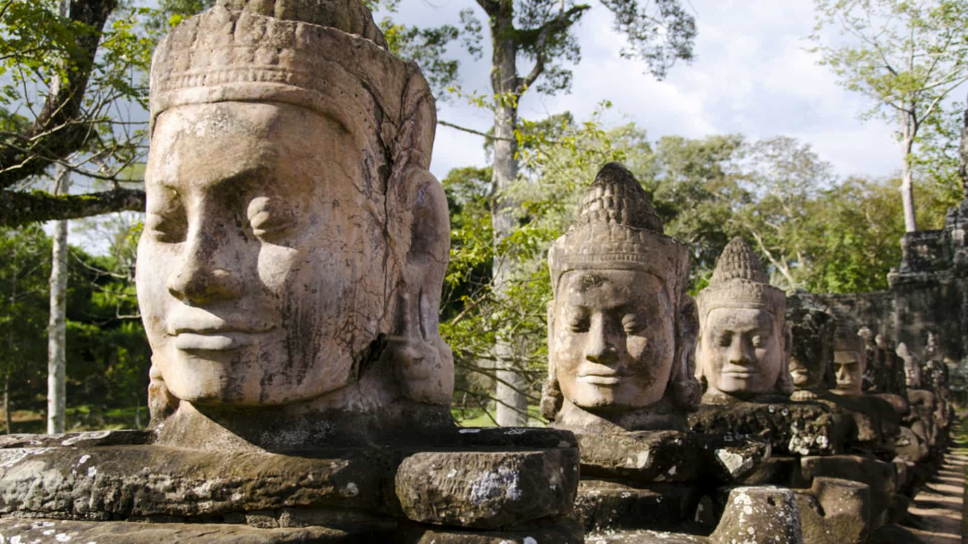 A row of ancient stone statues carved with serene facial expressions lining a bridge at the Angkor Thom causeway in Cambodia surrounded by trees.