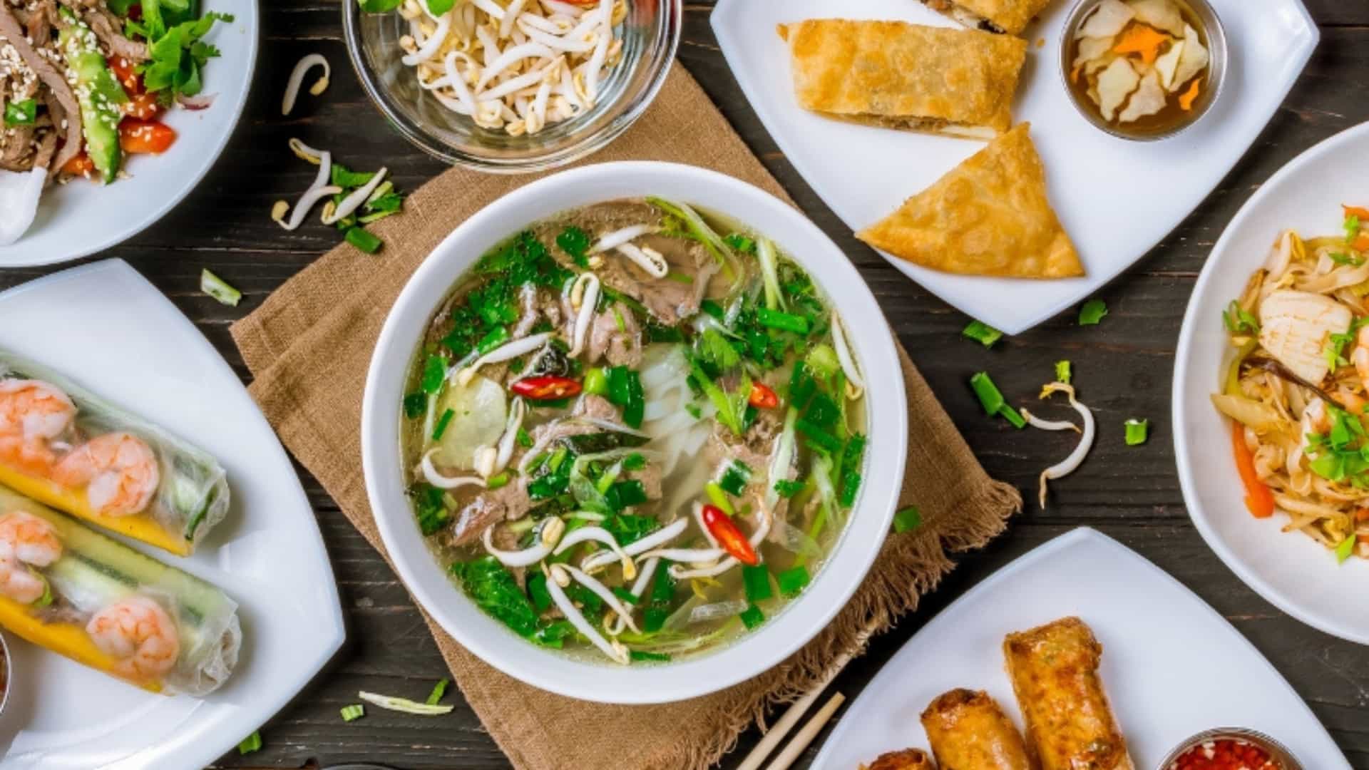 An overhead shot of a table covered with a variety of delicious Vietnamese dishes, representing the cuisine of the Southeast Asian river region. The centerpiece is a steaming bowl of beef pho with rice noodles, bean sprouts, and herbs. Around it are several plates holding different items, including fresh spring rolls filled with shrimp and vegetables, crispy fried spring rolls, and what appears to be a beef salad and pad thai. The food is arranged neatly on white plates, creating a colorful and appealing spread that highlights the rich culinary traditions of the region.