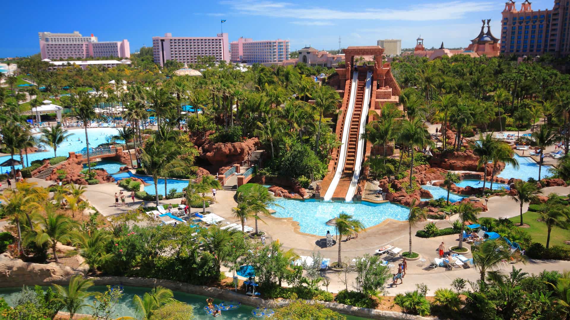 An aerial view of the massive water park at the Atlantis Resort on Paradise Island in the Bahamas. The central focus is a large, Mayan-themed temple structure with multiple water slides, including a steep drop slide and two parallel slides. People are visible on the slides and in the pools below. The water park is surrounded by winding lazy rivers, multiple pools, and lush palm trees. The large, pink-colored buildings of the resort stand in the background against a clear, blue sky, showcasing a popular tourist destination in the Bahamas.