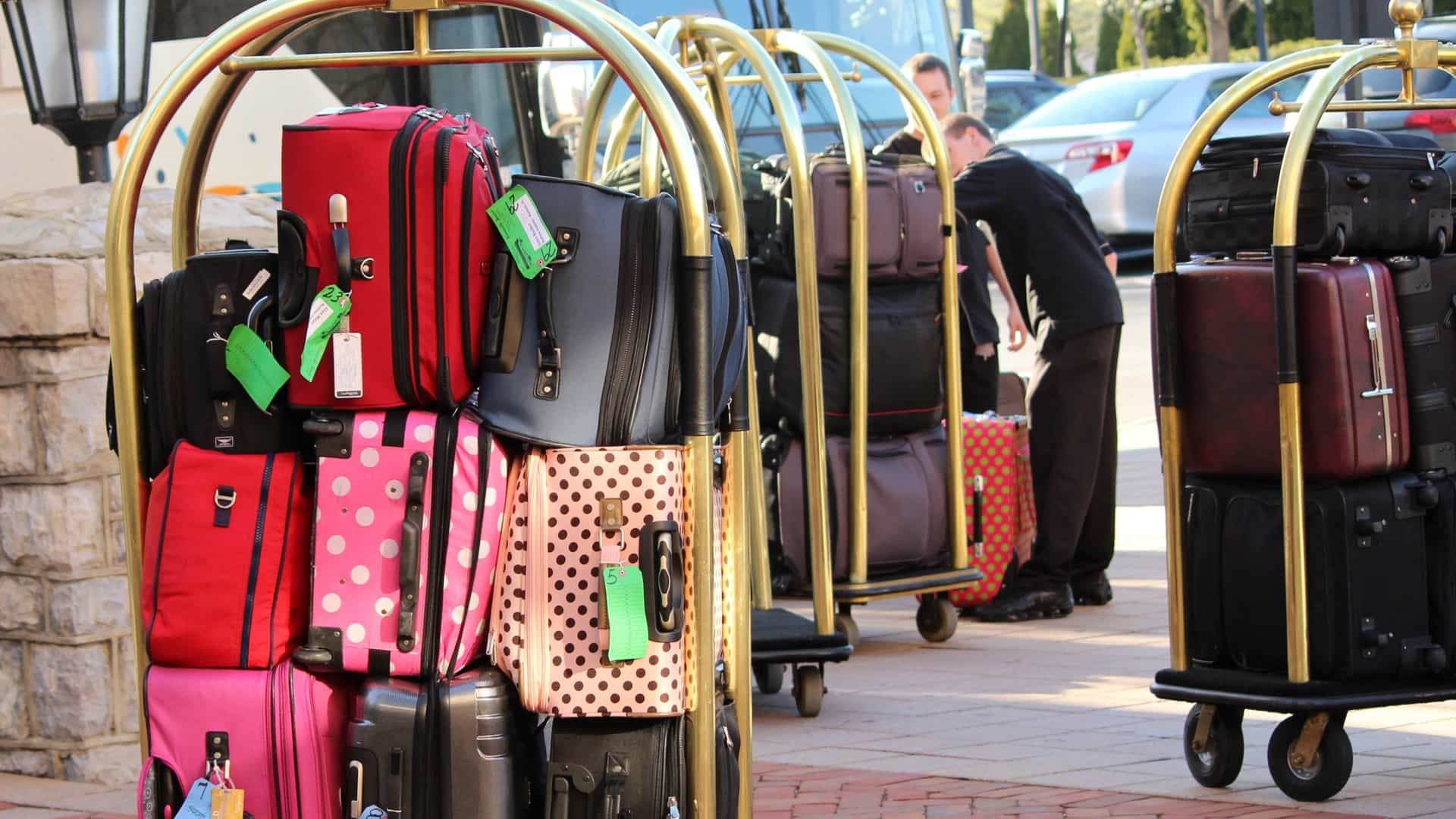 alt="Brass hotel luggage carts stacked high with colorful suitcases outside near waiting transport.