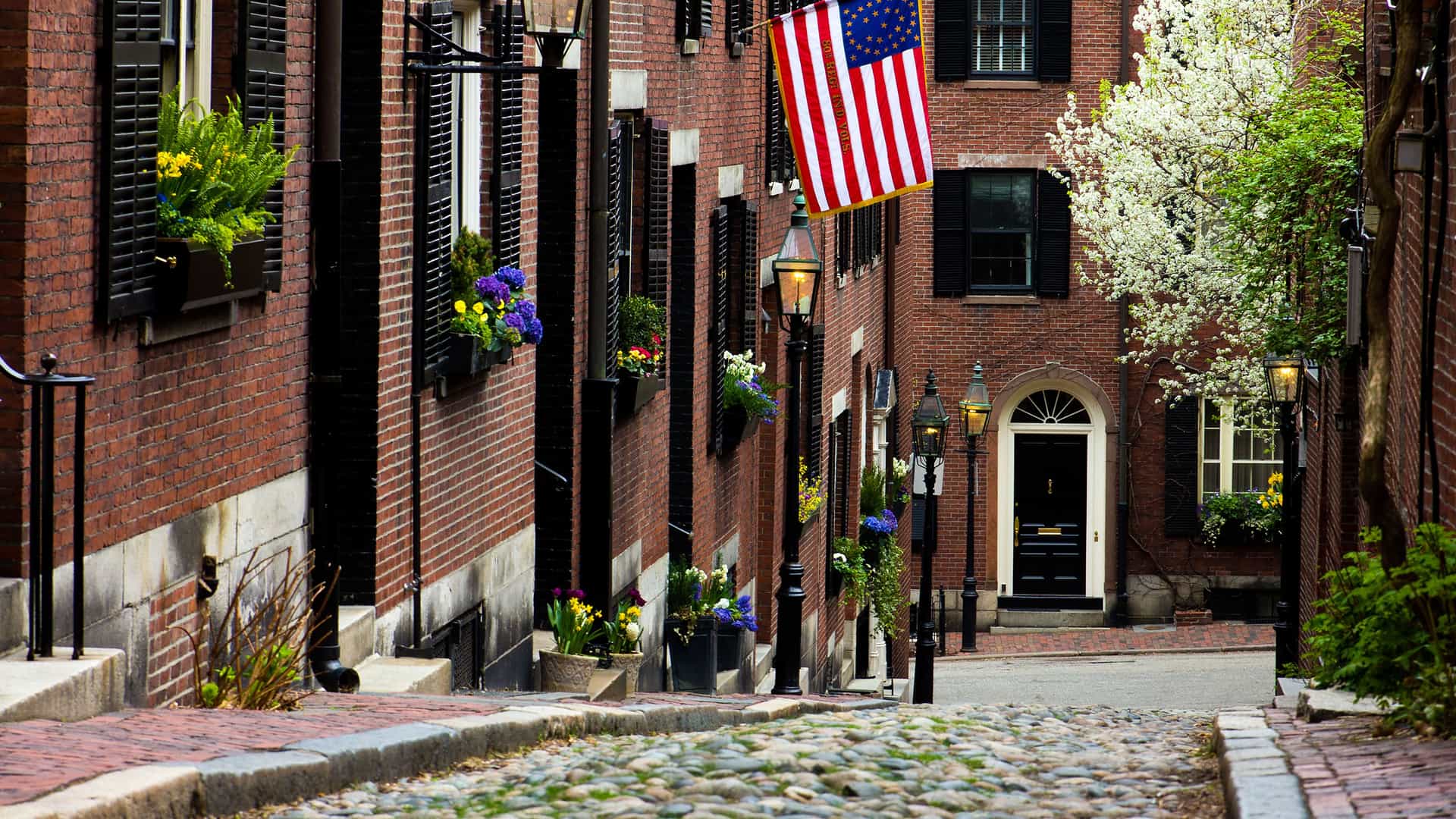 A charming, historic cobblestone street in the Beacon Hill neighborhood of Boston, a key city in the New England and Canada region. The street is lined with classic red brick townhouses featuring black shutters and window boxes overflowing with colorful flowers. An American flag with a circle of stars hangs from one of the buildings. Gas-style street lamps and a blooming white tree add to the picturesque scene, which captures the old-world charm and colonial history of Boston.