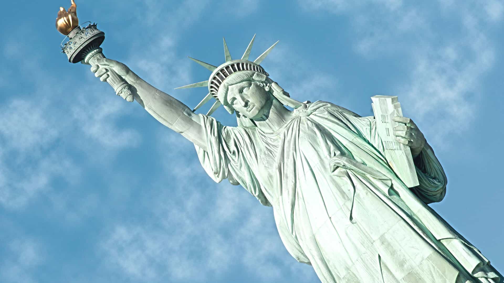An upward-angled close-up of the Statue of Liberty in New York. The statue's weathered green copper surface is visible, showing its flowing robes and spiked crown. She holds a golden torch high in her right hand and a tablet in her left. The background is a vibrant blue sky with soft, white clouds. The image highlights the detailed features of this famous American symbol and landmark in the Canada and New England region.