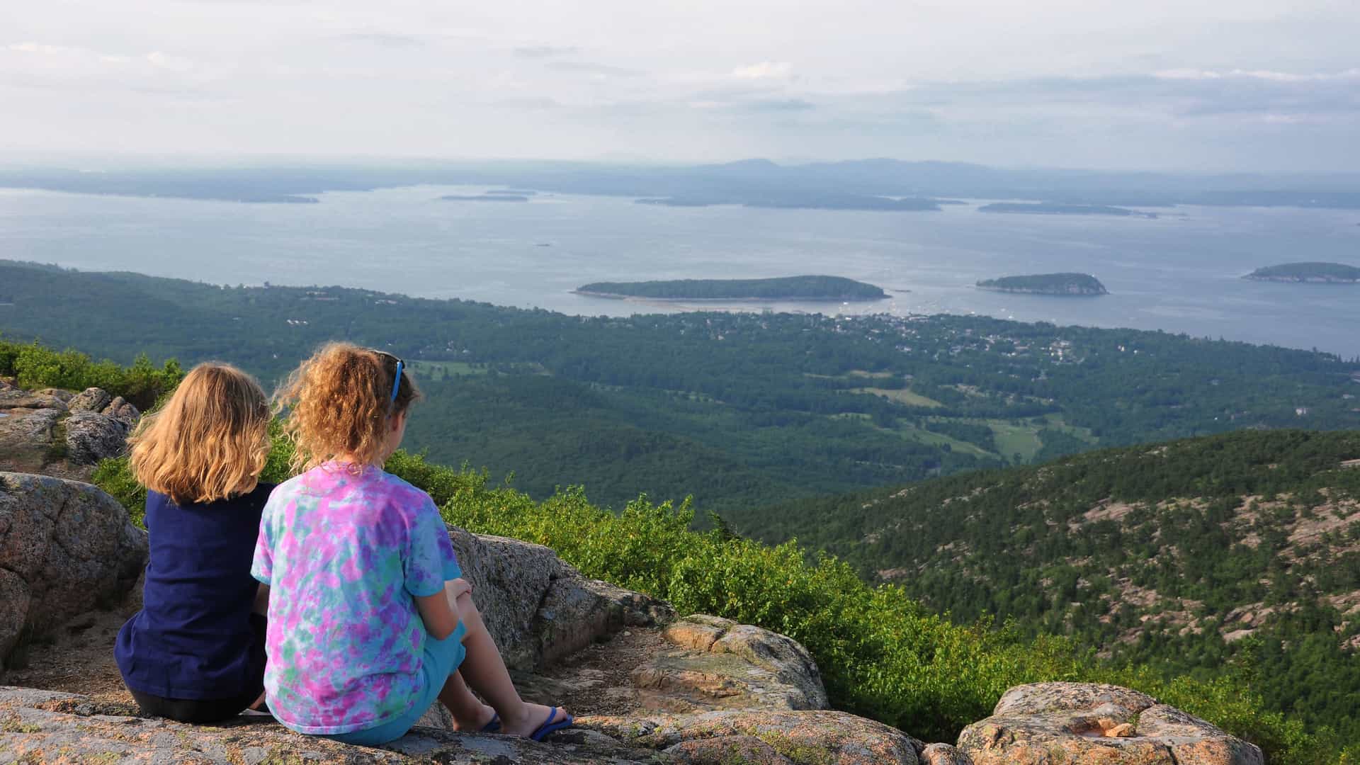Two young girls are sitting on a rocky outcrop on Cadillac Mountain in Acadia National Park, Maine, and looking out at the panoramic view. The girl on the left has long, blonde hair and is wearing a dark blue shirt. The girl on the right is wearing a colorful, tie-dyed shirt. Below them, a vast landscape of rolling, green hills and dense forests extends to the coastline. The calm, gray-blue water of the ocean is dotted with numerous small islands. The hazy sky above adds to the serene and expansive scene, representing a key natural landscape of the New England and Canada region.