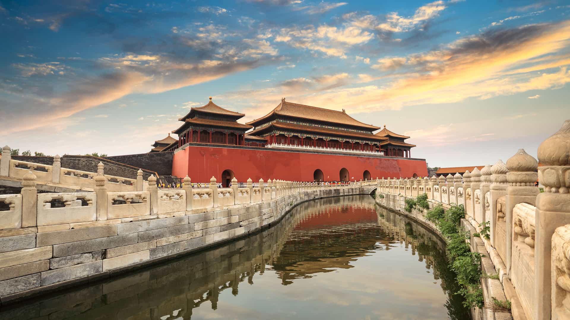 The iconic red walls and golden roofs of the Forbidden City in Beijing, China, are beautifully reflected in the calm waters of the surrounding moat. A traditional stone bridge with ornate carvings crosses the water, leading toward the grand palace buildings. The sky above is a vibrant mix of blue, orange, and white clouds during sunset. The image captures the historical grandeur and imperial architecture of this significant landmark in China.