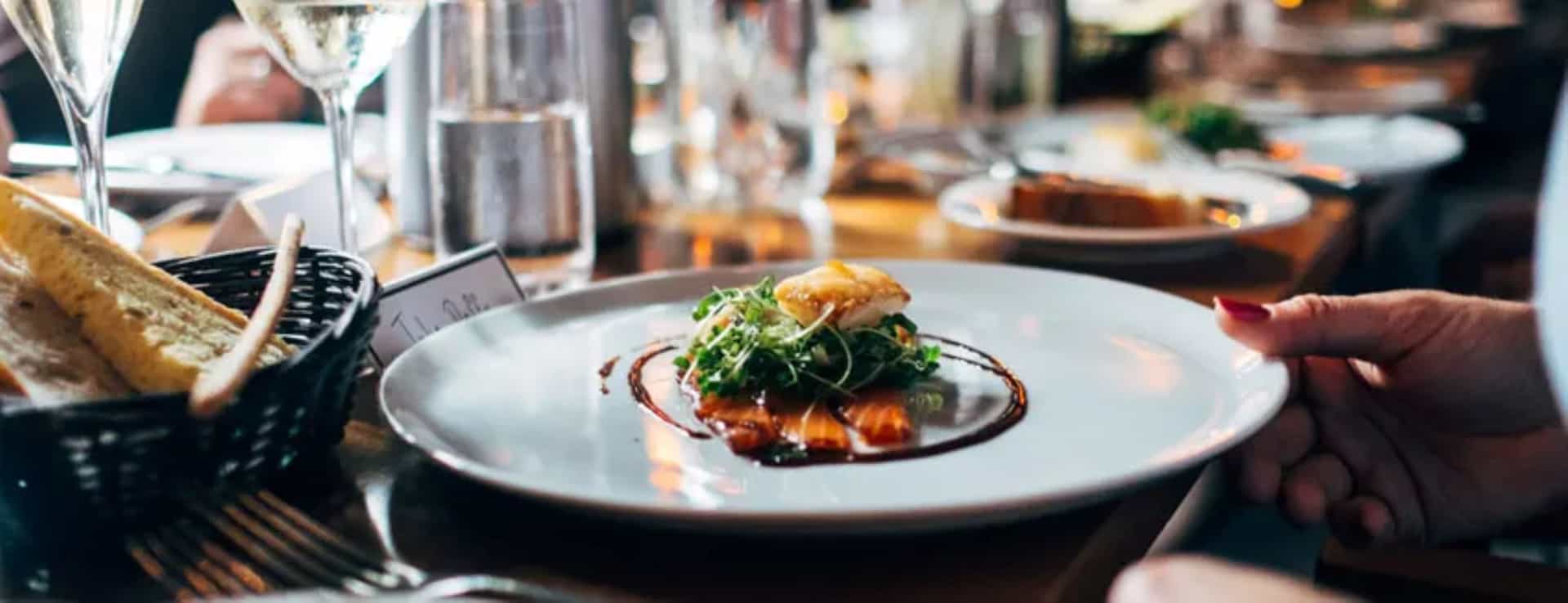 A plate of fine dining cuisine being served next to a bread basket on a table with glasses and other plates.