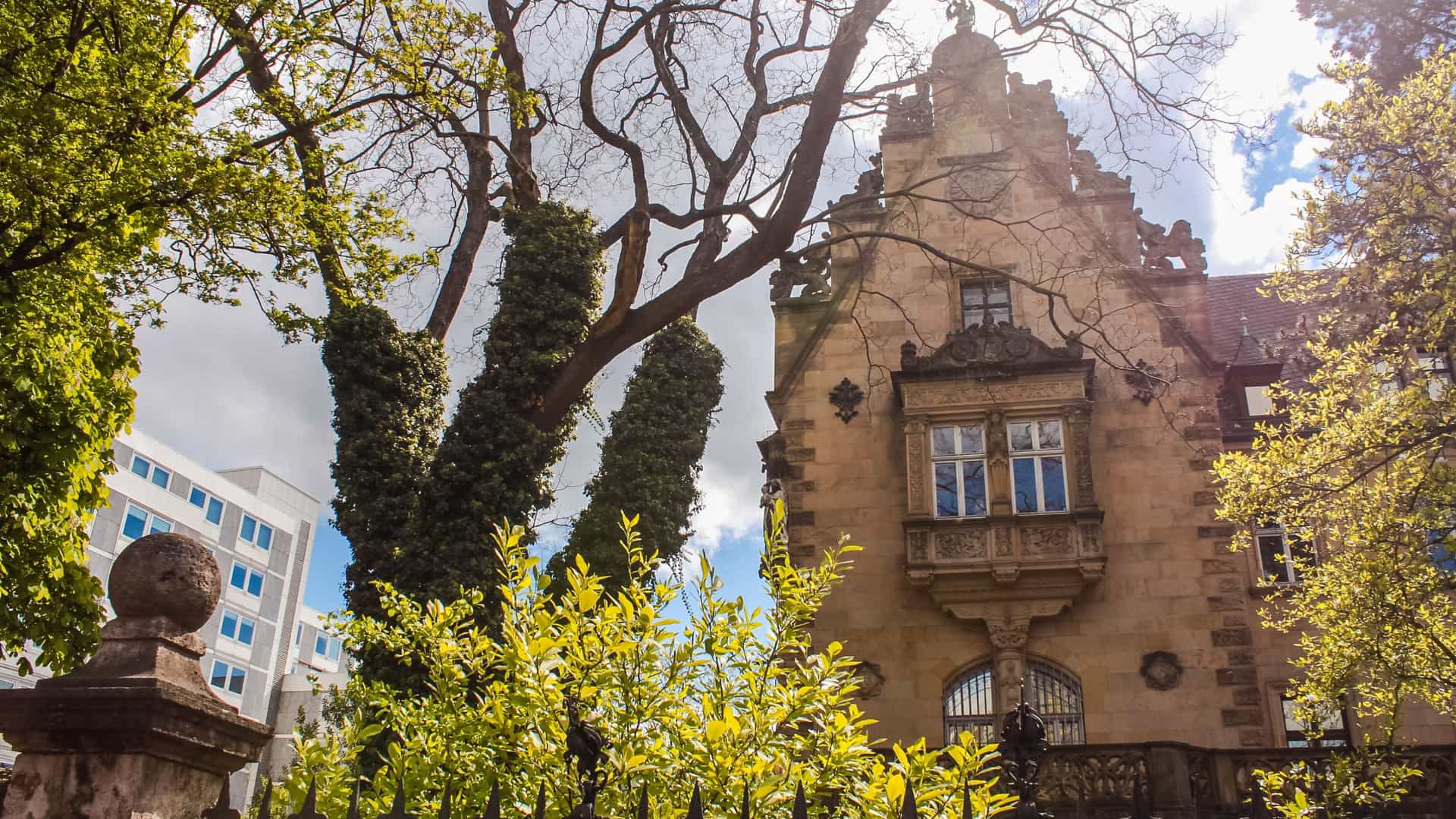 The image displays the stately exterior of the Liebieghaus Skulpturensammlung (Sculpture Collection), a museum housed in a 19th-century villa in Frankfurt am Main, Germany. The building is surrounded by lush green trees.
