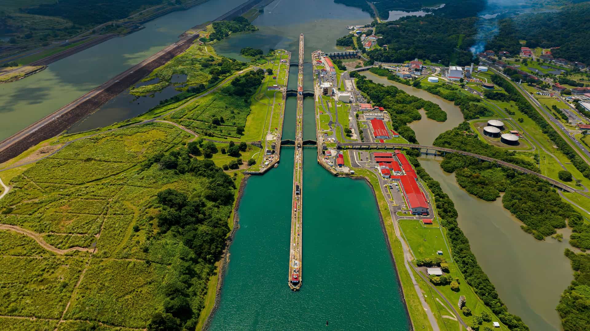 An aerial view of the Panama Canal, showing a massive container ship transiting the locks, surrounded by dense jungle and infrastructure.