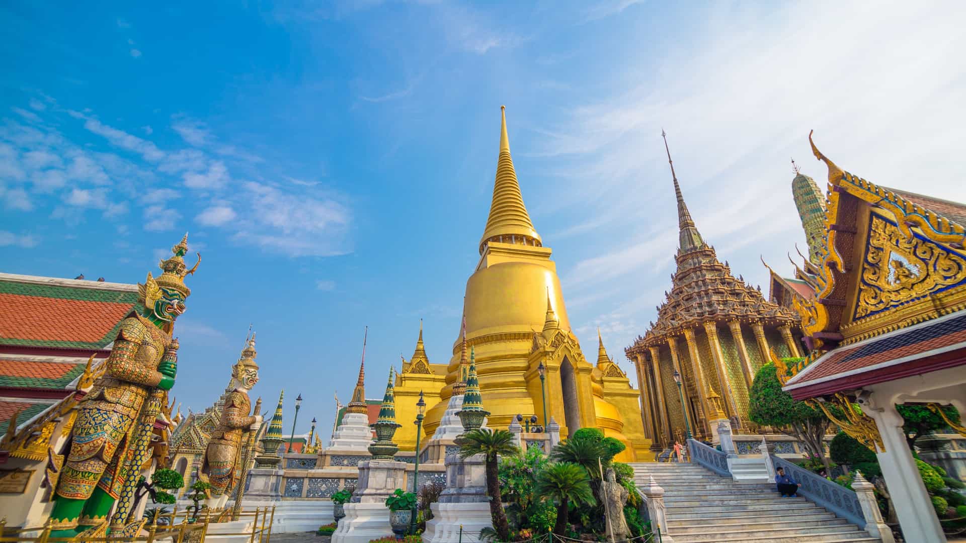 A close-up view of the Wat Phra Kaew (Temple of the Emerald Buddha) complex in Bangkok, Thailand, showing a golden chedi (stupa) and a building with a multi-tiered roof, both decorated with intricate mosaics and carvings. The structures are surrounded by green trees and are set against a clear blue sky.