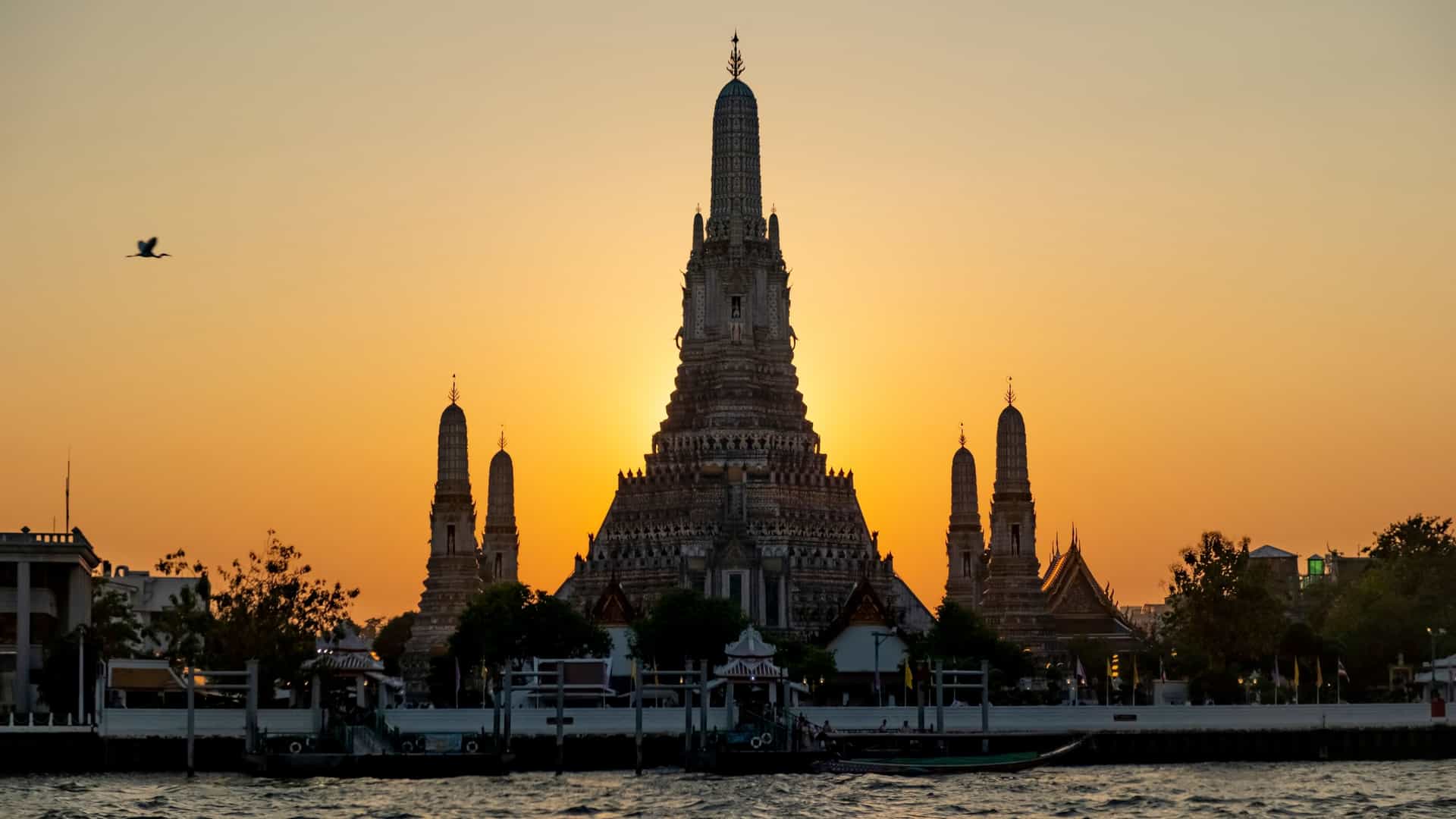 A scenic view of the Wat Arun temple in Bangkok, Thailand, located on the banks of the Chao Phraya River. The central prang (spire) and surrounding smaller spires, decorated with colorful porcelain and seashells, rise majestically against a bright sky.