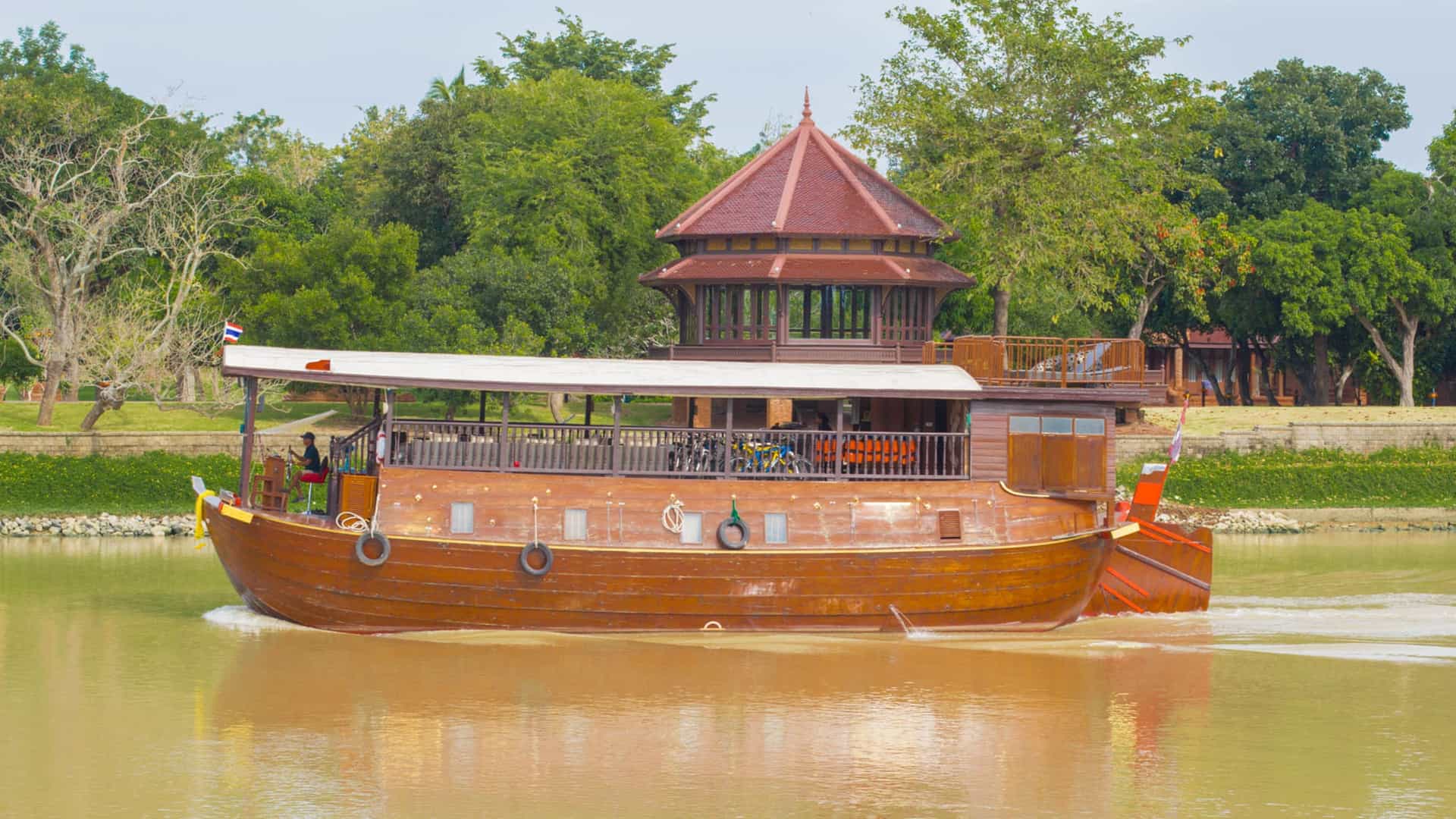 A large, multi-deck dinner cruise ship sailing on the Chao Phraya River in Bangkok, Thailand, with the illuminated cityscape and Thai temples, such as Wat Arun or The Grand Palace, visible along the riverbanks, likely at night or twilight.