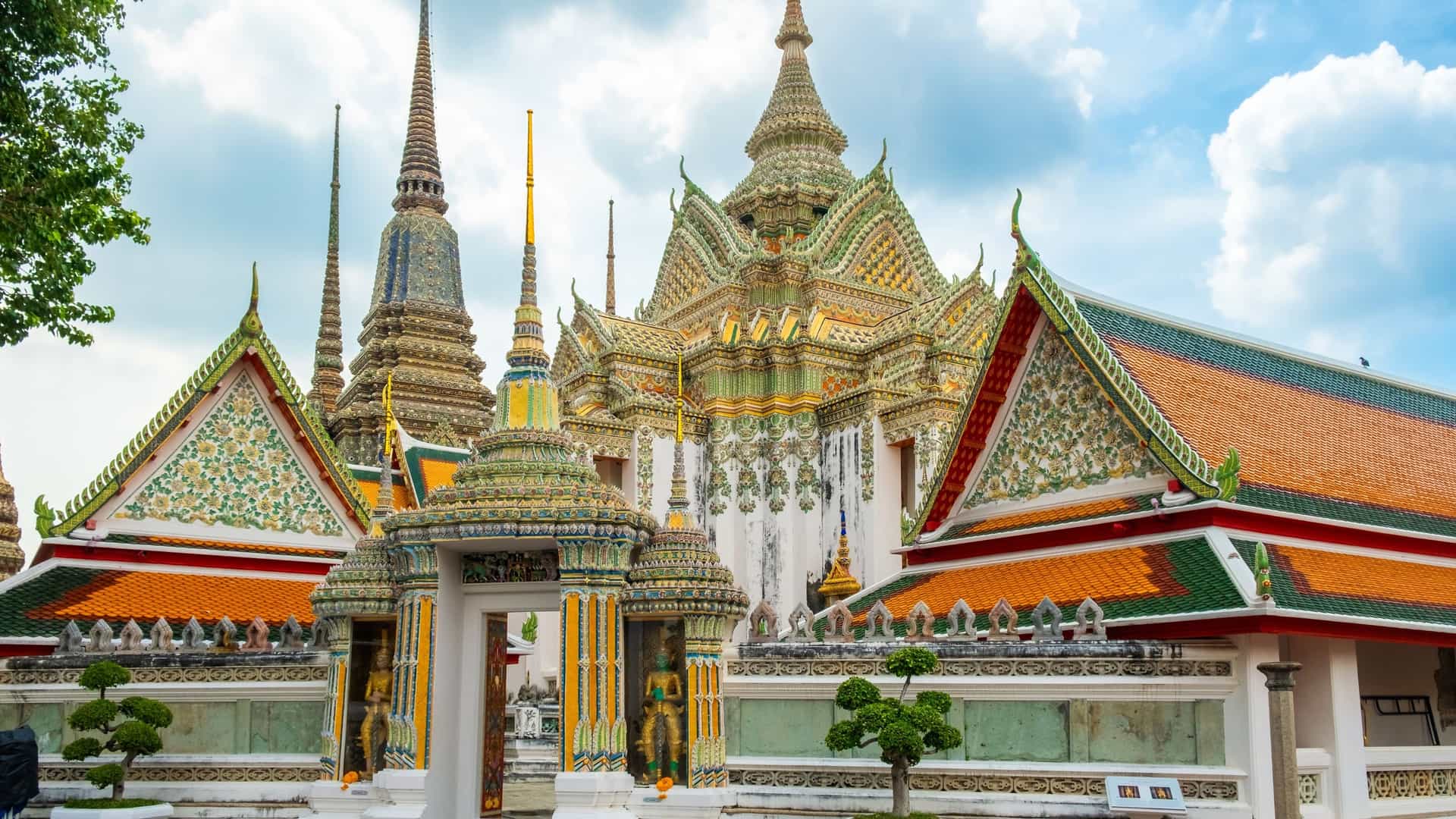 A scenic view of the Wat Pho temple complex in Bangkok, Thailand, featuring several ornate, multi-colored chedis (stupas) and pavilions with tiered roofs. The structures are intricately decorated with ceramic mosaics and are set within a courtyard surrounded by trees.
