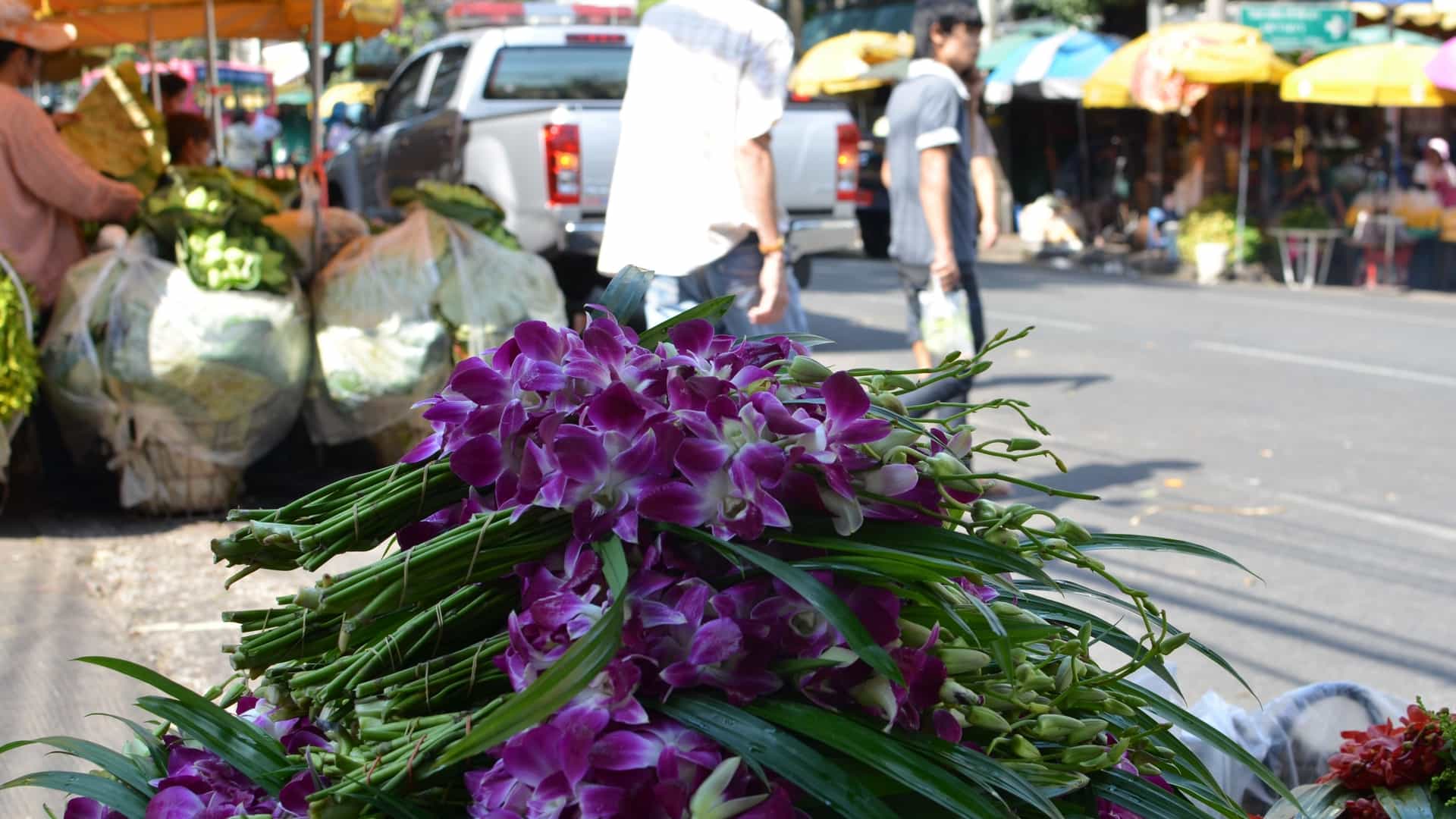 A close-up view of vibrant purple orchid bundles with long green stems, likely for sale, at the bustling Pak Khlong Talat Flower Market in Thailand. In the background, out of focus, market stalls with yellow umbrellas, people, and a white pickup truck are visible on a sunny day.