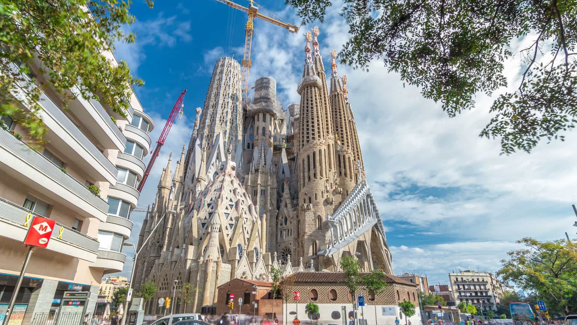 A scenic view of the iconic Sagrada Familia Roman Catholic Church in Barcelona, Spain. The basilica's towering, intricate spires dominate the skyline, contrasting with the vibrant scene of lush green autumn trees in the foreground and a dramatic, blue cloudy sky overhead. The light and colors suggest a daytime, clear-weather setting.
