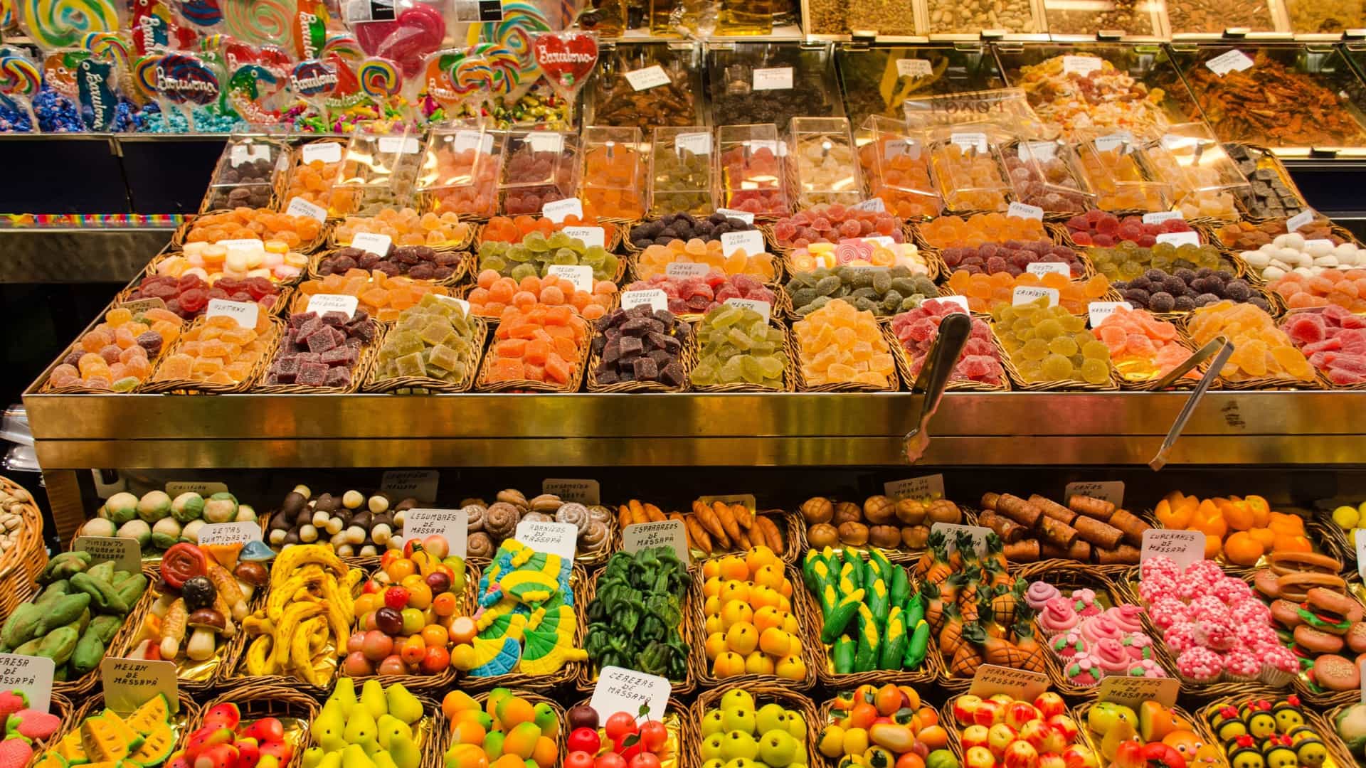 A vibrant, close-up view of a stall at La Boqueria Food Market in Barcelona, Spain, featuring a dense display of colorful sweets. The candies, including various types of fruit jellies or marzipan, are arranged in large, tiered, stacked cones or mounds, showcasing a rainbow of colors like red, green, yellow, and orange, covered in a light dusting of sugar. The presentation is rich and tempting.