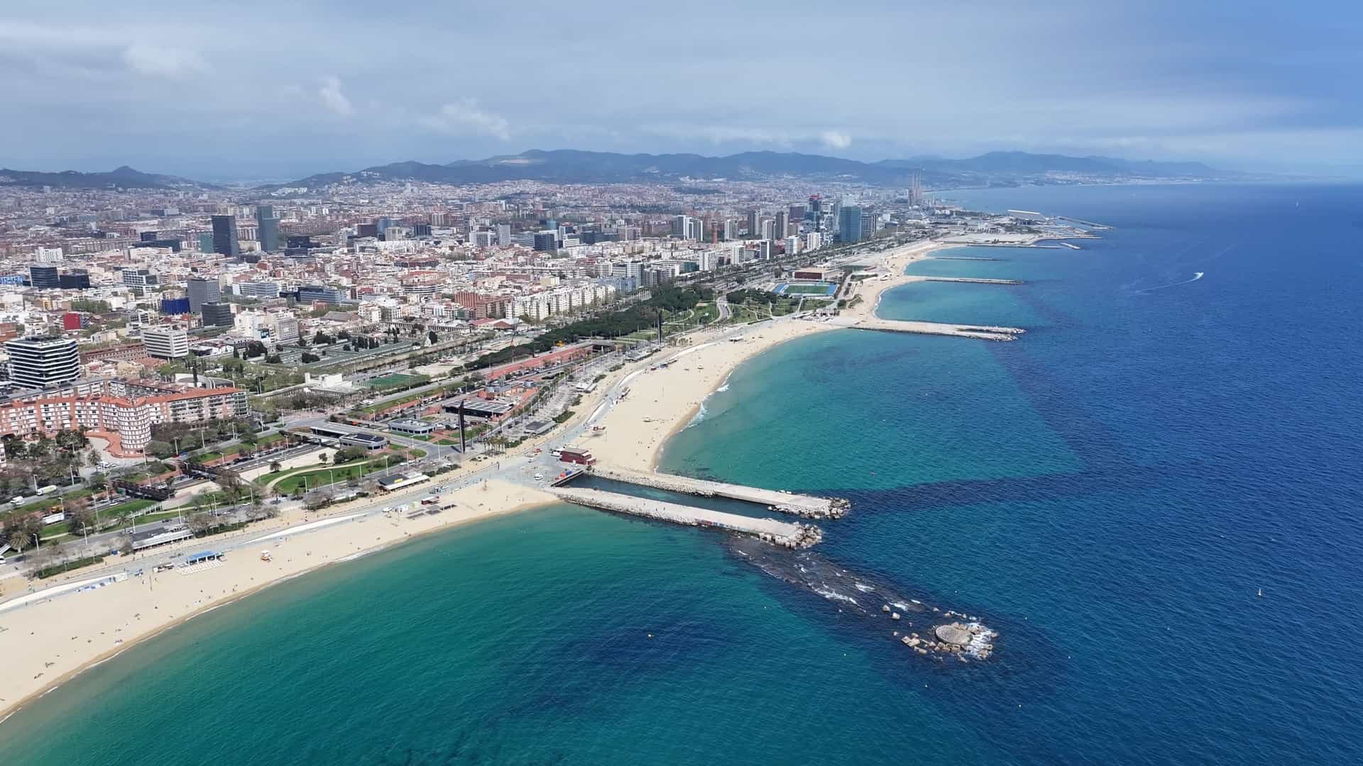A stunning cityscape view of Barceloneta Beach in Barcelona, Catalonia, Spain. The image features a wide expanse of sand with the Mediterranean Sea on one side and the city's high-rise buildings and skyline in the background.