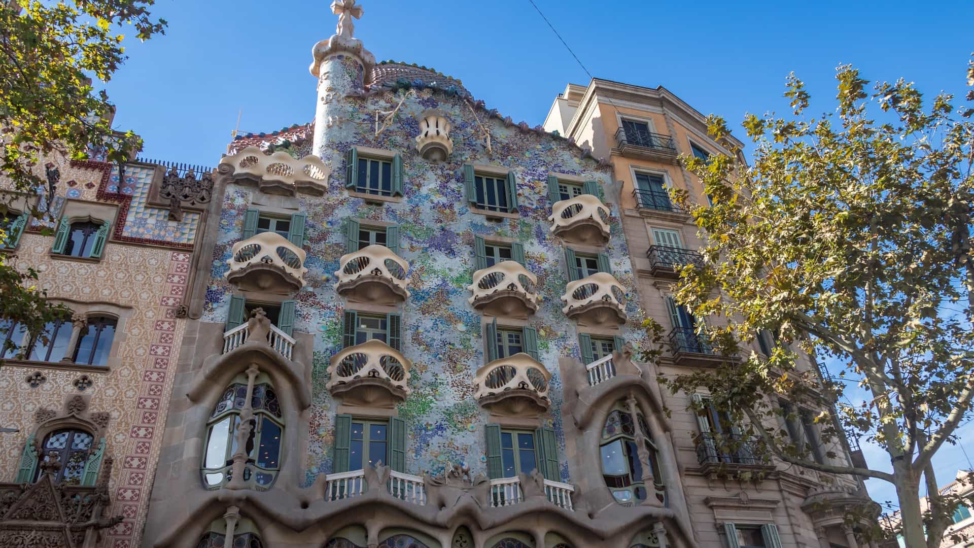 A bright, clear daytime view of the main facade of Casa Batlló in Barcelona, Spain, a famous modernist building by Antoni Gaudí. The facade features an intricate mosaic of colorful, broken ceramic tiles (trencadís) that shimmer under the sunlight. The building is characterized by its signature wave-like stonework, the elongated, bone-shaped columns, and the elliptical, mask-like balconies with wrought iron railings. The colorful, scaled roof and the turret topped with Gaudí's four-armed cross are visible against the blue sky.