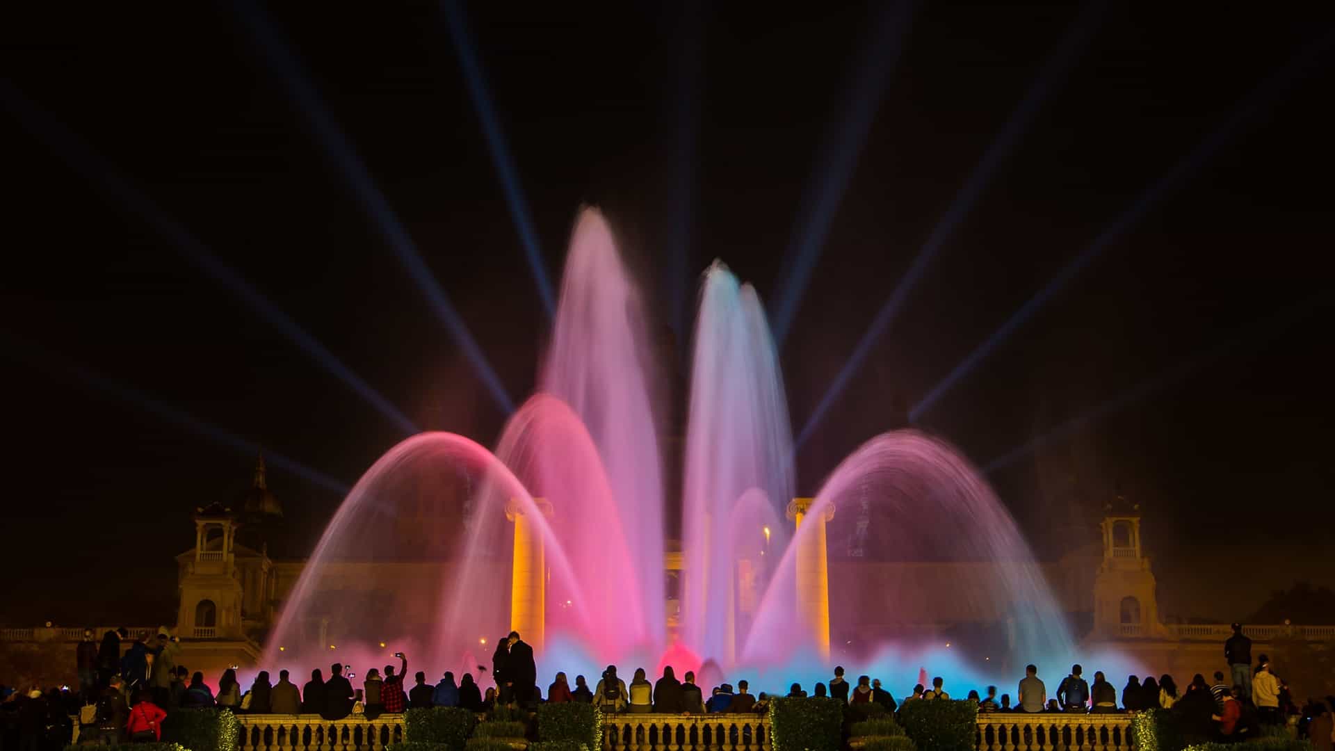 A nighttime view of the Magic Fountain of Montjuïc in Barcelona, Spain, during one of its famous light and music shows. The fountain's water jets are illuminated with vibrant colors against the dark night sky. The scene includes the silhouettes of people gathered to watch the spectacle.