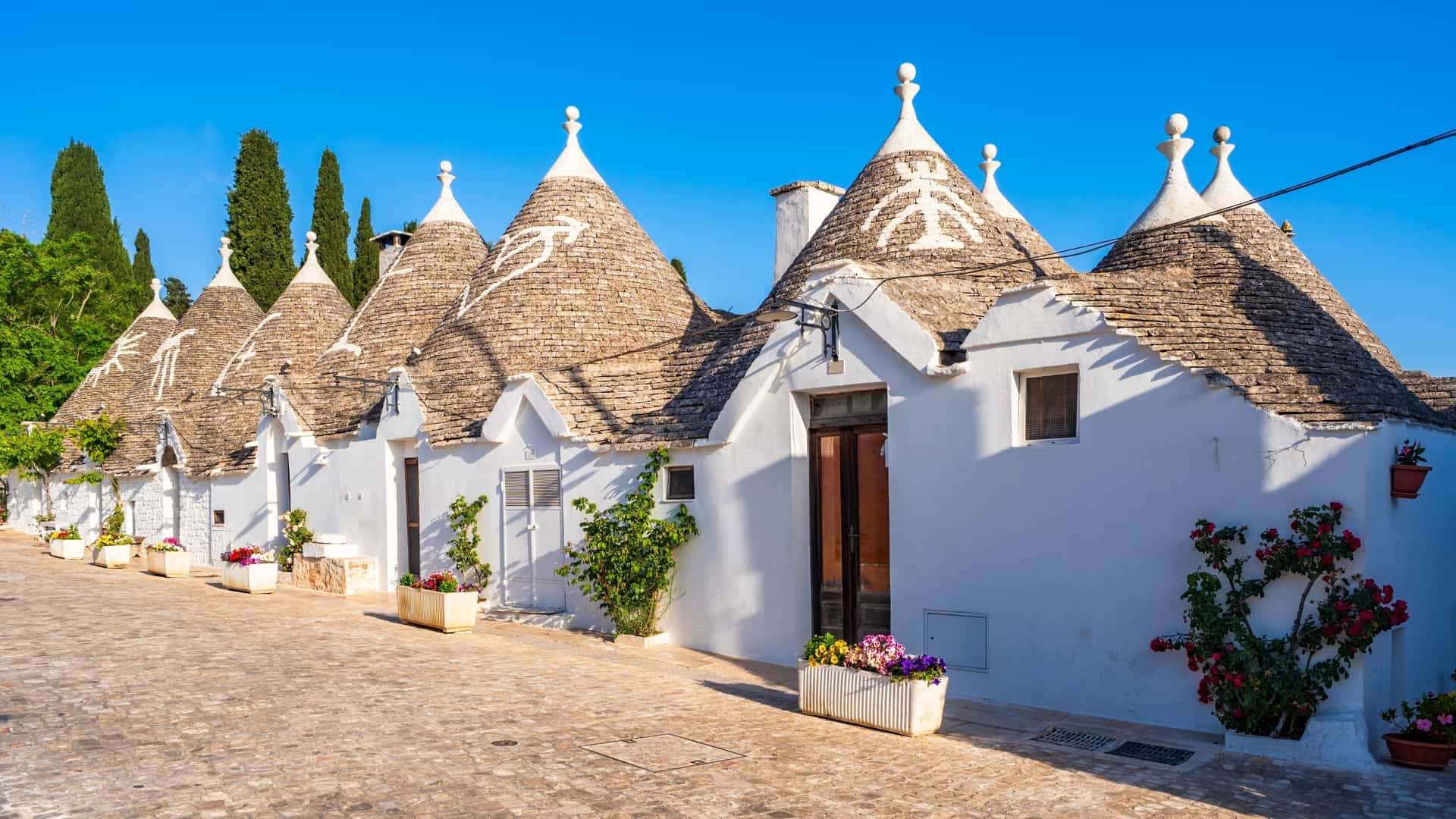 A street view of the famous historical Trulli houses in Puglia, Italy, featuring their distinctive white, dry-stone walls and conical gray roofs.