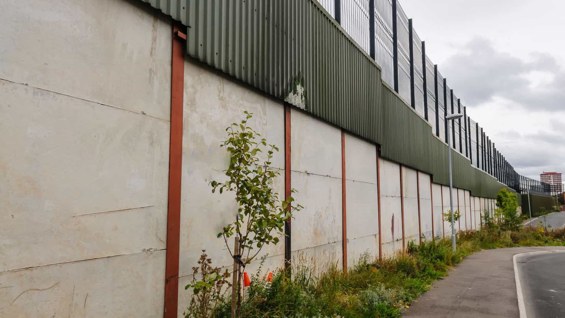  A tall, grey concrete Peace Wall in Belfast, Northern Ireland, covered in various colorful murals and messages. A metal fence and barbed wire sit atop the wall. The art features political and international solidarity themes, reflecting the divided communities the wall separates. A black taxi, often used for political tours, is visible near the wall.