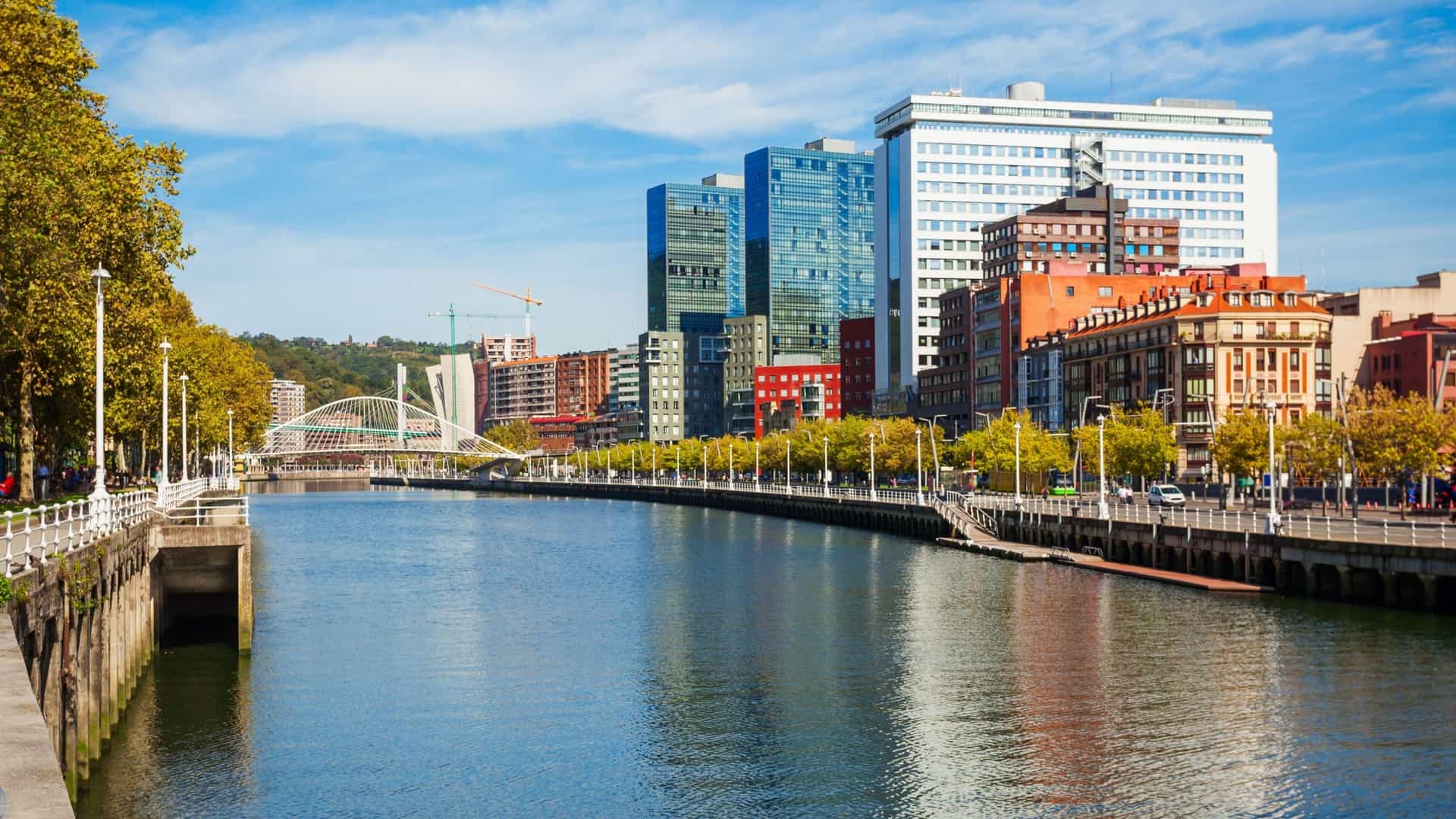  An aerial view of the Nervión River and its embankment in Bilbao, Spain. The river flows through the city center, with a wide, developed walking path and modern architecture visible along the banks. The image captures the city's successful urban regeneration, with the river serving as a central feature.