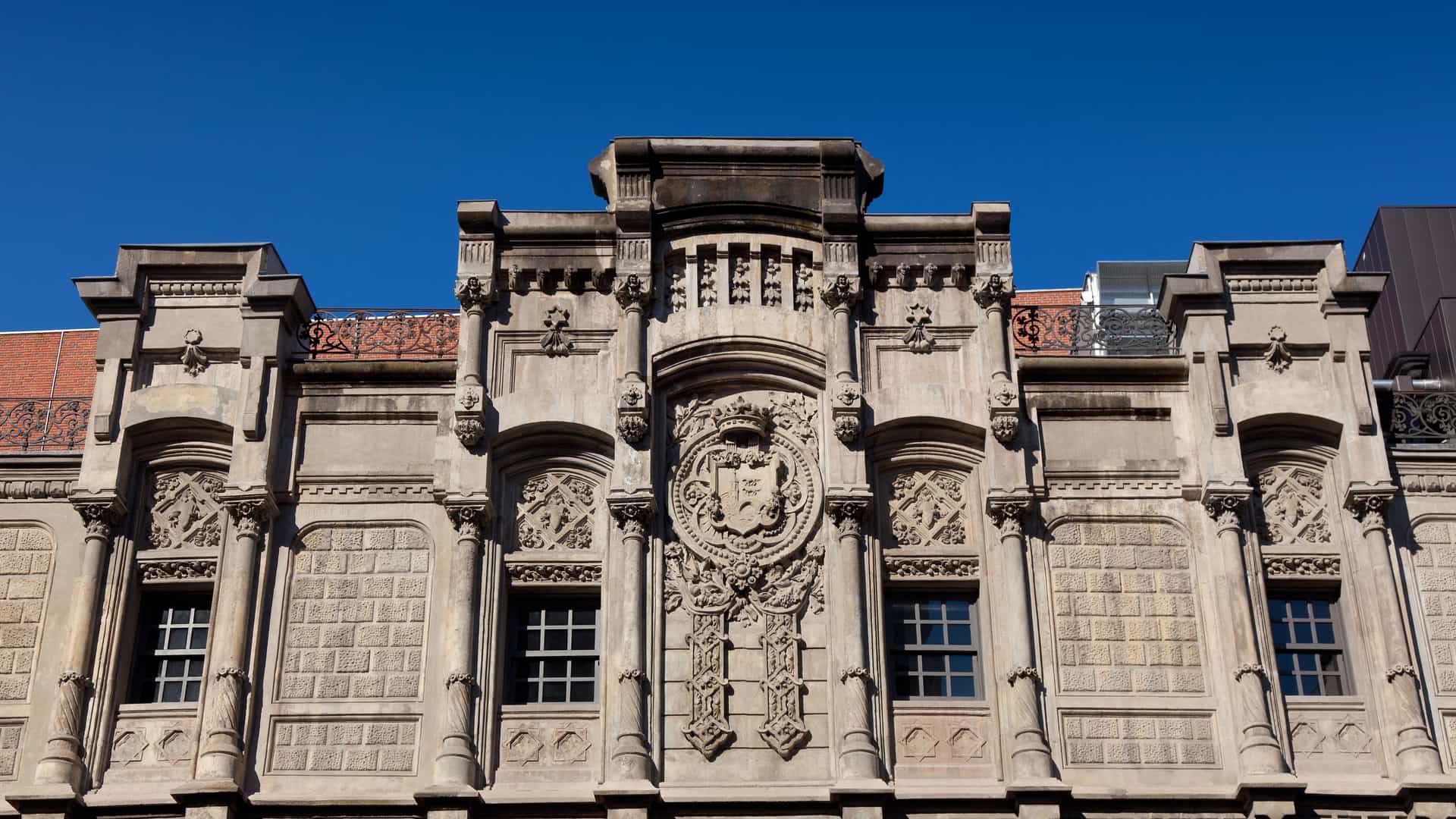  The facade of Azkuna Zentroa (formerly Alhóndiga Bilbao), a large, historic building in Bilbao, Spain. The architecture is a distinctive early 20th century modernist or eclectic style, featuring a brick and stone exterior with numerous symmetrically arranged windows and decorative trim. The building has a tower at the corner and is a prominent cultural and leisure center in the city.