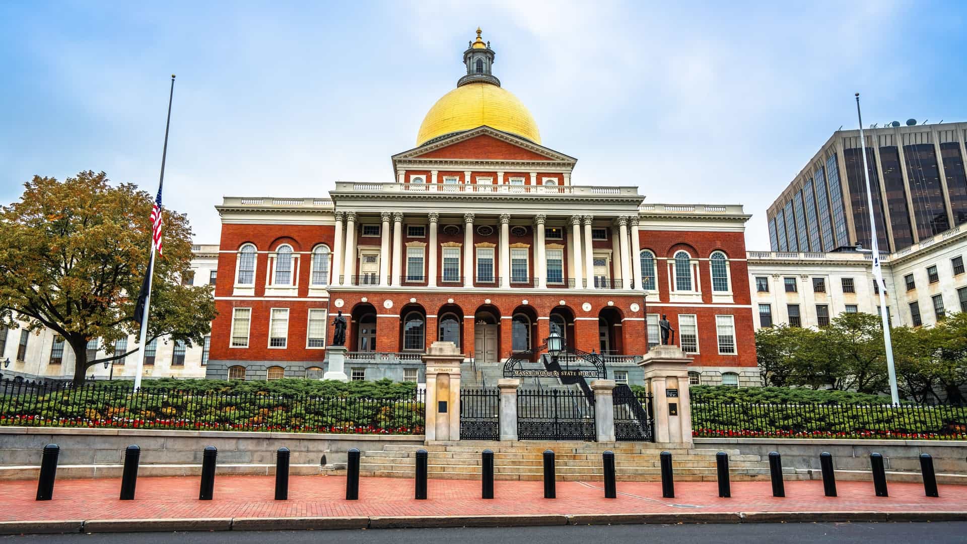 The gold-domed Massachusetts State House in Boston is viewed from a street lined with historical architecture and parked cars.