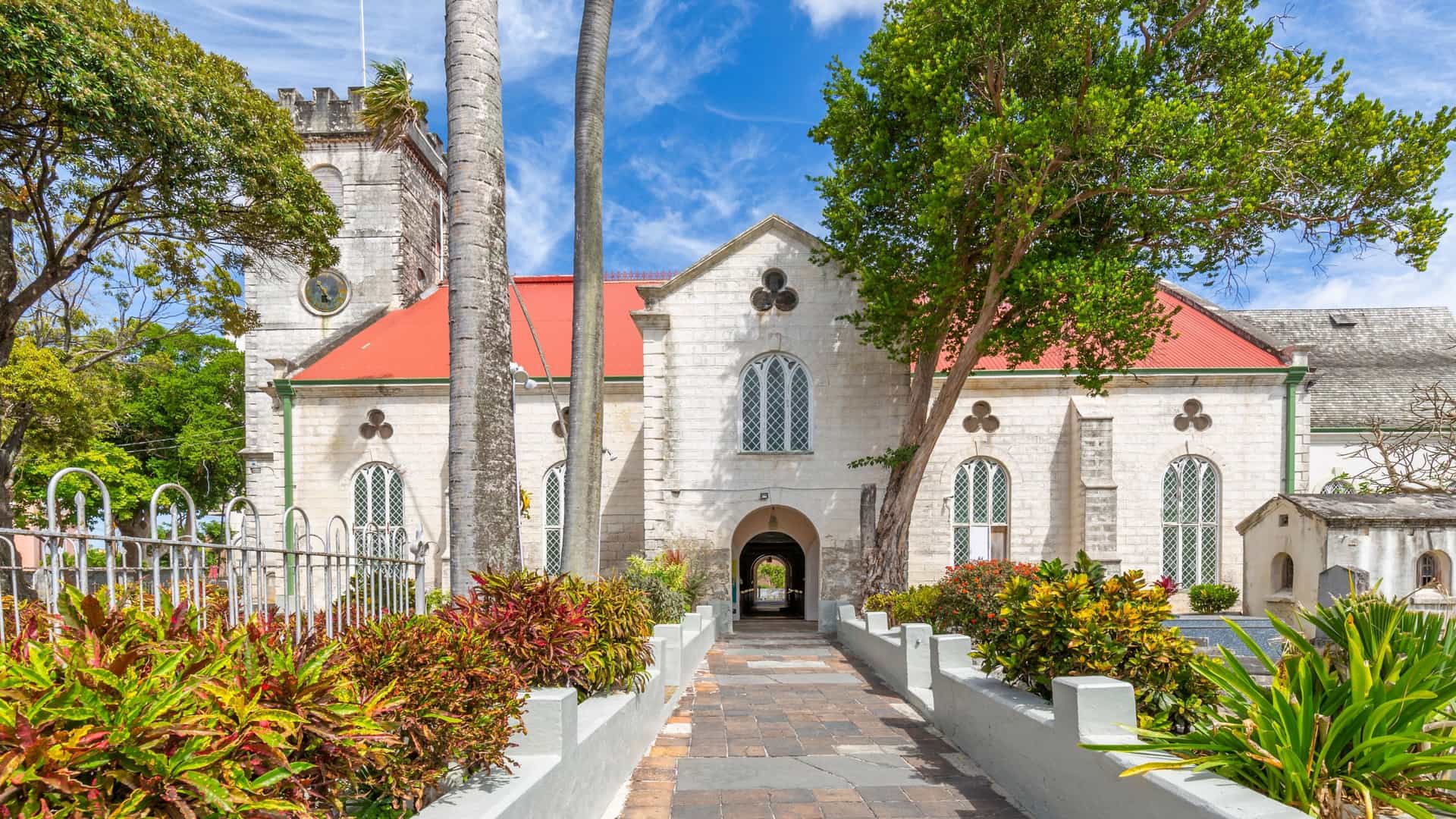  An exterior view of the Cathedral Church of Saint Michael and All Angels in Bridgetown, Barbados. The historic building, constructed of light colored coral stone, features a prominent central tower and is surrounded by lush green tropical foliage and trees.