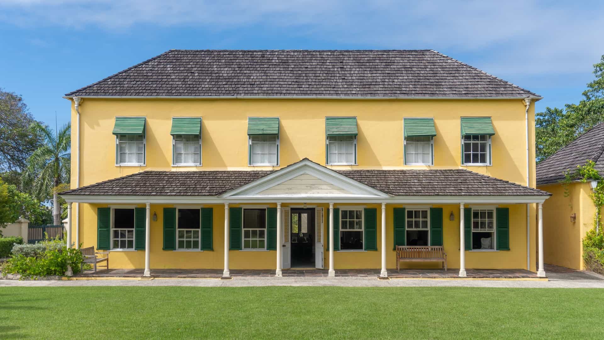  The George Washington House in Bridgetown, Barbados. This historic, Georgian style mansion is a two story building with a light colored exterior, a red or dark colored hipped roof, and a gallery or balcony on the upper level, typical of 18th century colonial architecture. It is situated within the Garrison Historic Area, often surrounded by a well maintained lawn and tropical landscaping.