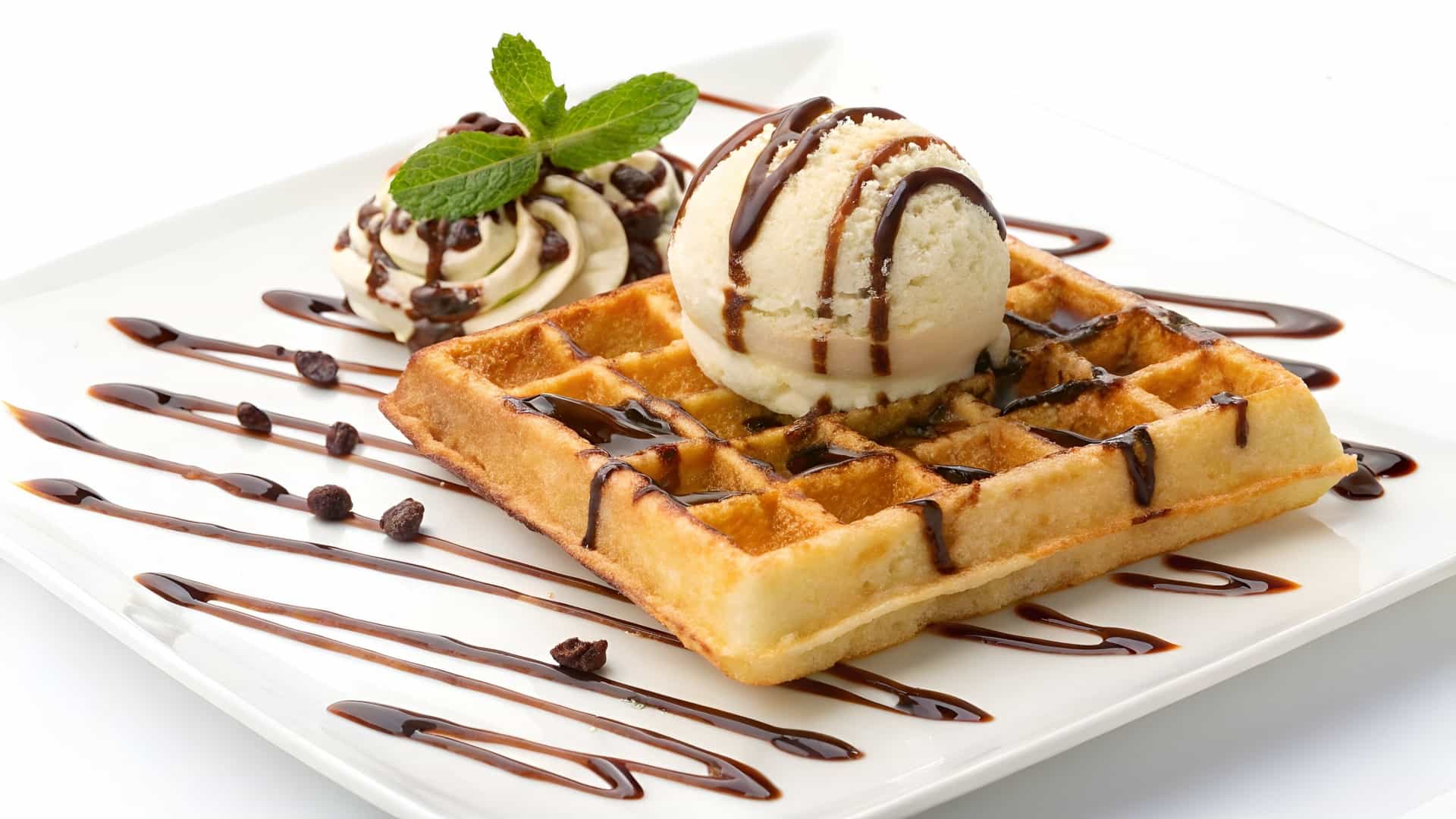 A top-down view of a thick, square-shaped Belgian waffle topped with a scoop of white ice cream, sliced fresh strawberries, and a drizzle of sauce, presented on a round plate.