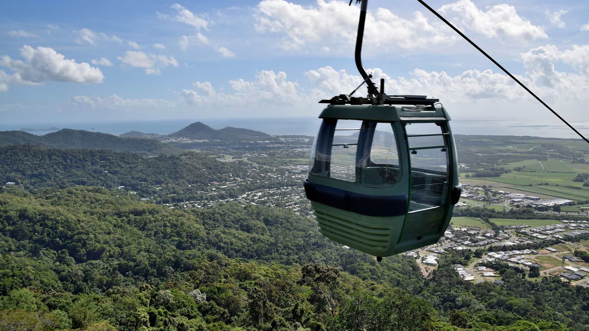  An expansive aerial view of a cable car (gondola) transporting passengers high above the dense, lush green canopy of a tropical rainforest. The image, likely depicting the Skyrail Rainforest Cableway in Cairns, Northern Queensland, Australia, shows a gondola cabin zipping along a wire with the mountainous, unbroken expanse of the forest stretching out to the horizon under a bright sky.