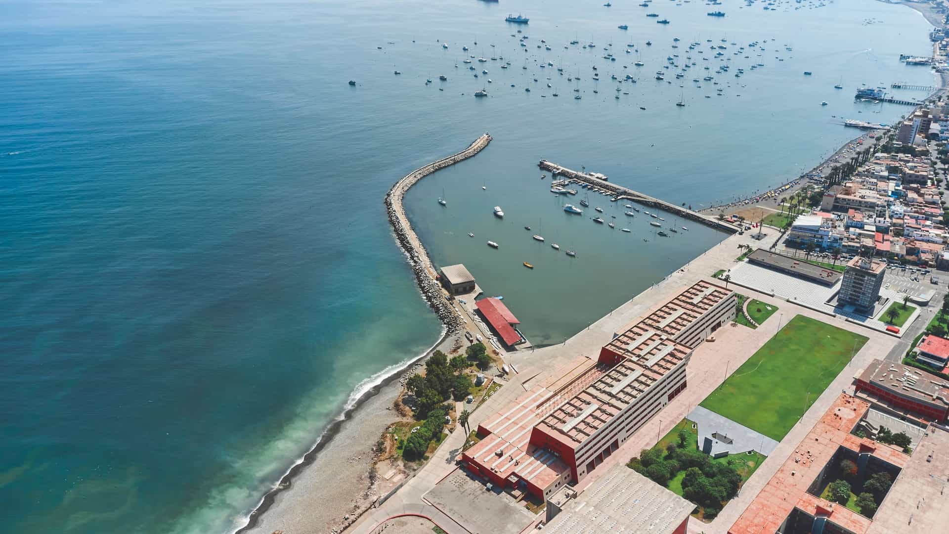A high-angle aerial, panoramic view of the La Punta district in Callao, Peru, which extends into the Pacific Ocean like a peninsula. The image shows the distinct grid pattern of the residential area, characterized by its colorful houses, surrounded on three sides by the deep blue ocean, with the long, curving coastline visible in the background.