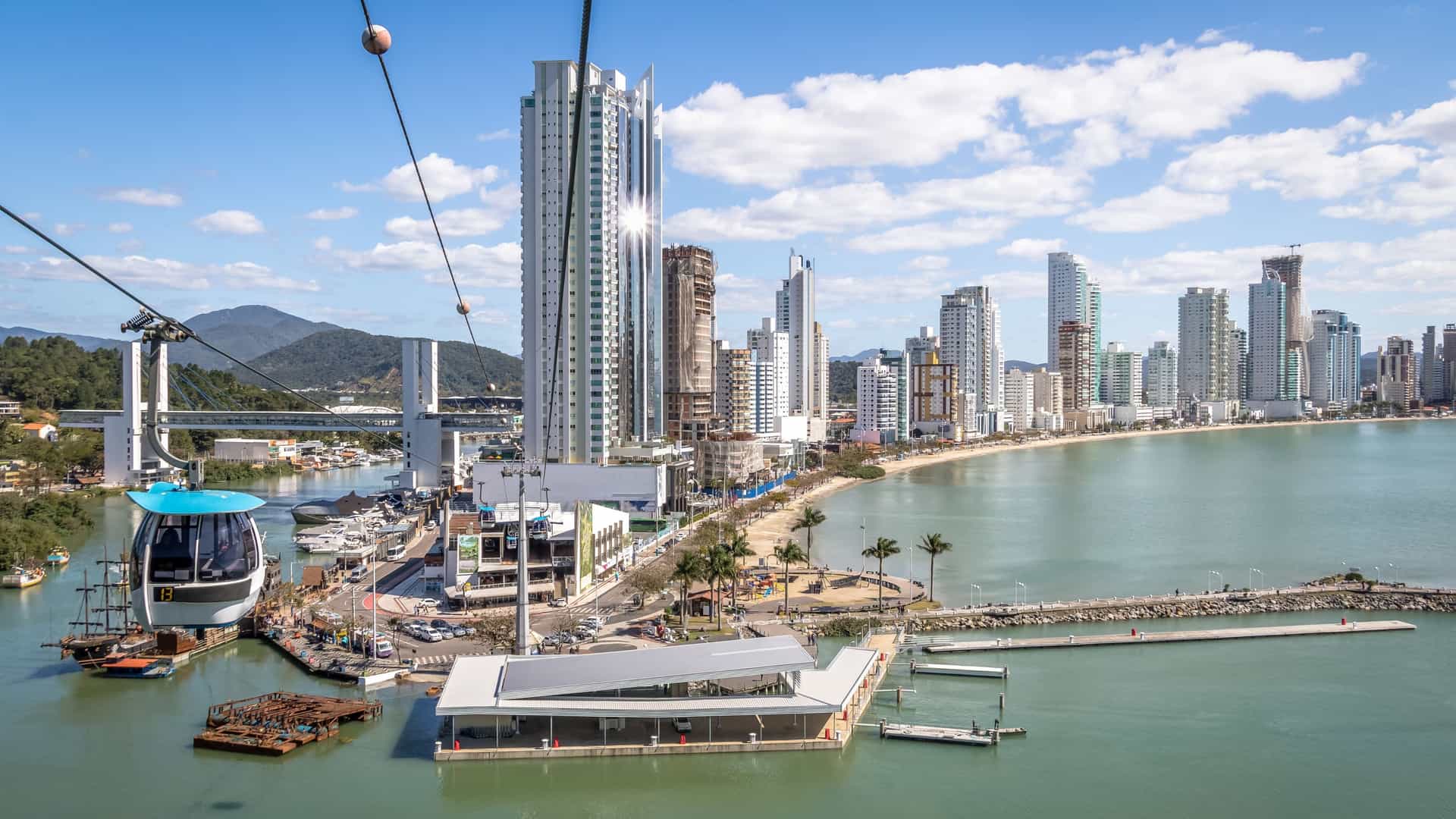 An aerial view of Balneário Camboriú, Santa Catarina, Brazil, showing the densely packed high-rise buildings along the curved coastline, with the wide beach and the ocean. The image also captures a gondola lift (cable car) rising up the hillside toward the Christ Light statue (Cristo Luz).