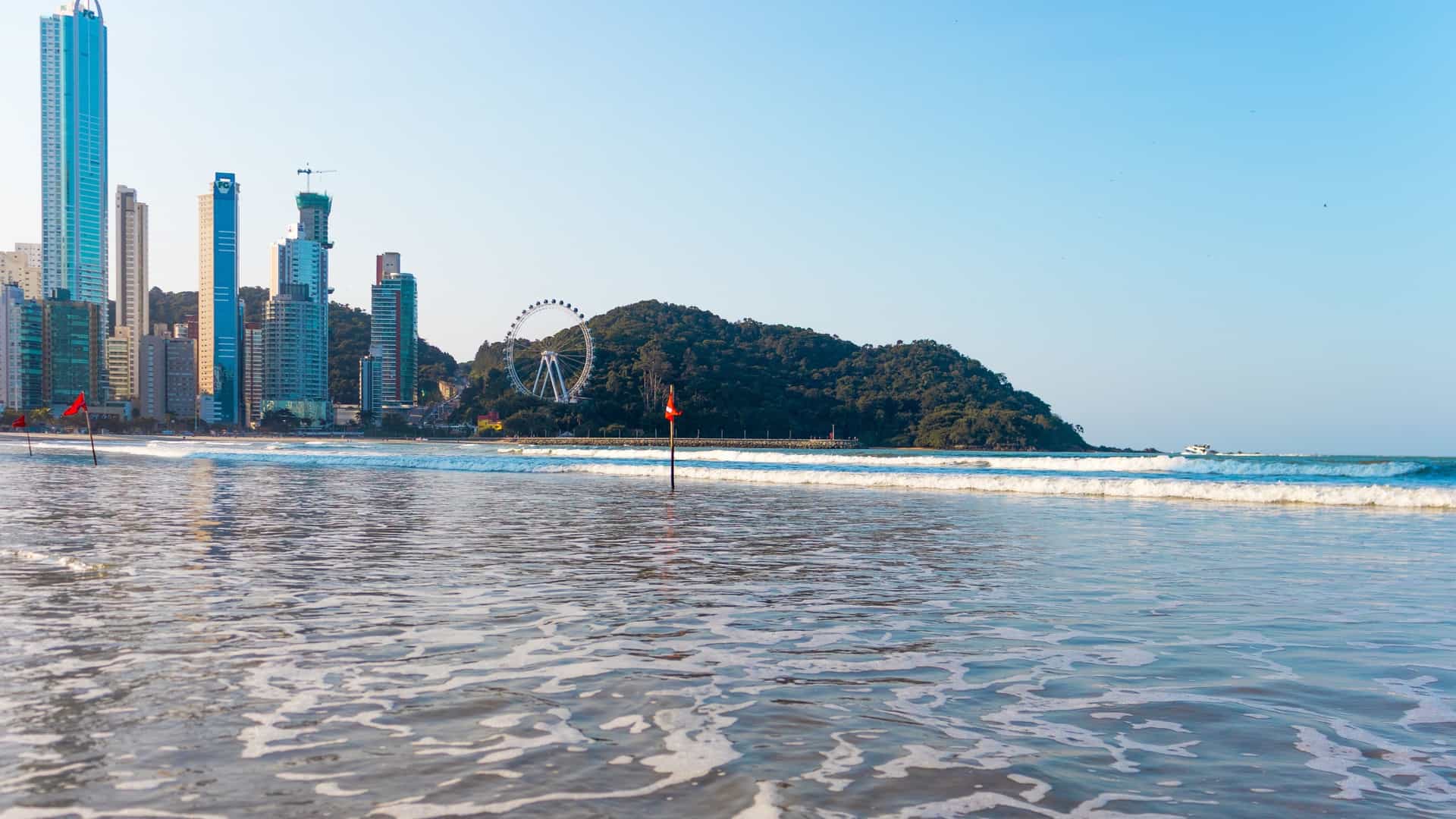 An aerial view of the dense, highly developed coastline of Balneário Camboriú, Santa Catarina, Brazil, showing a wide stretch of sandy beach and the Atlantic Ocean framed by towering high-rise buildings.