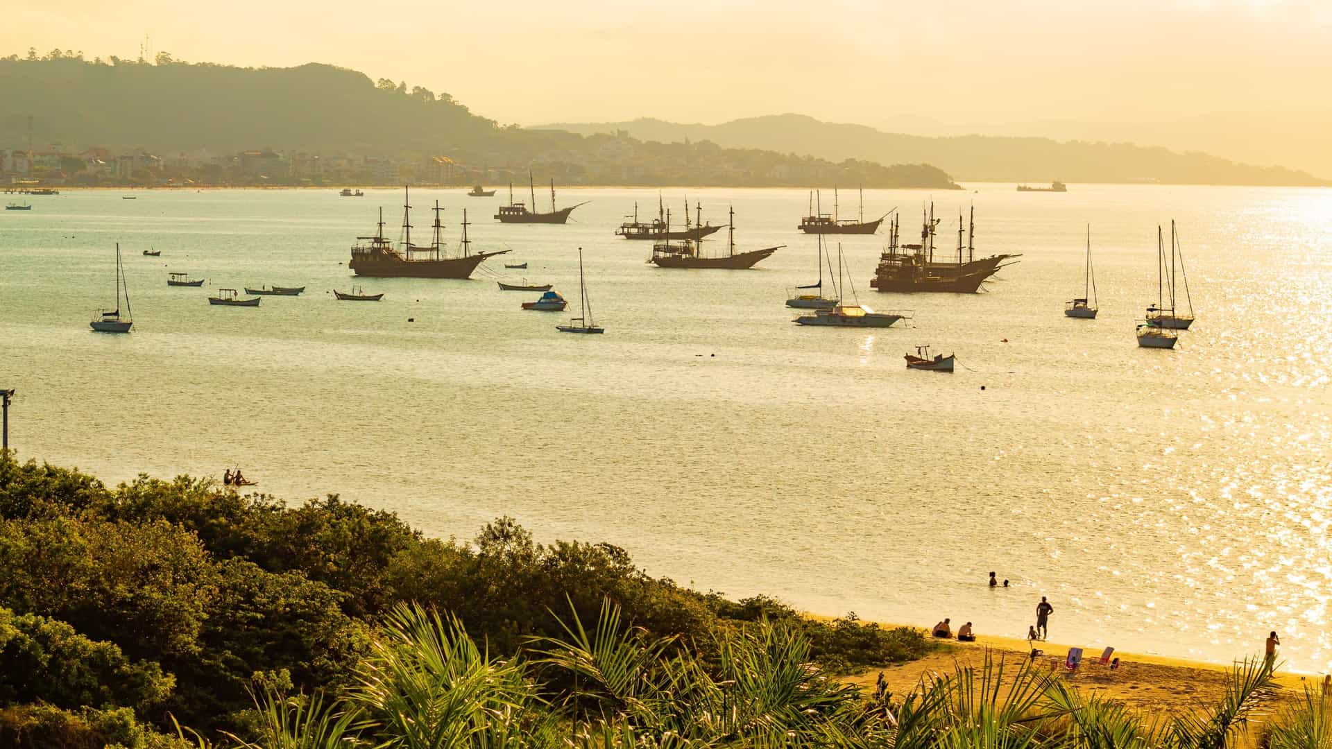 A group of brightly painted, pirate-themed tour boats (escunas piratas) are docked in a small bay, waiting for passengers. The bay is located near the Cachoeira do Bom Jesus beach, which is a popular departure point for these themed tours in Florianópolis, Santa Catarina, Brazil.