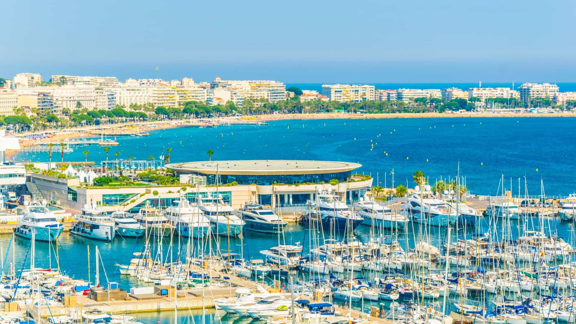 An aerial view of Cannes, France, showing the city's dense urban area, the curving coastline of the French Riviera, and the harbor filled with yachts and boats.