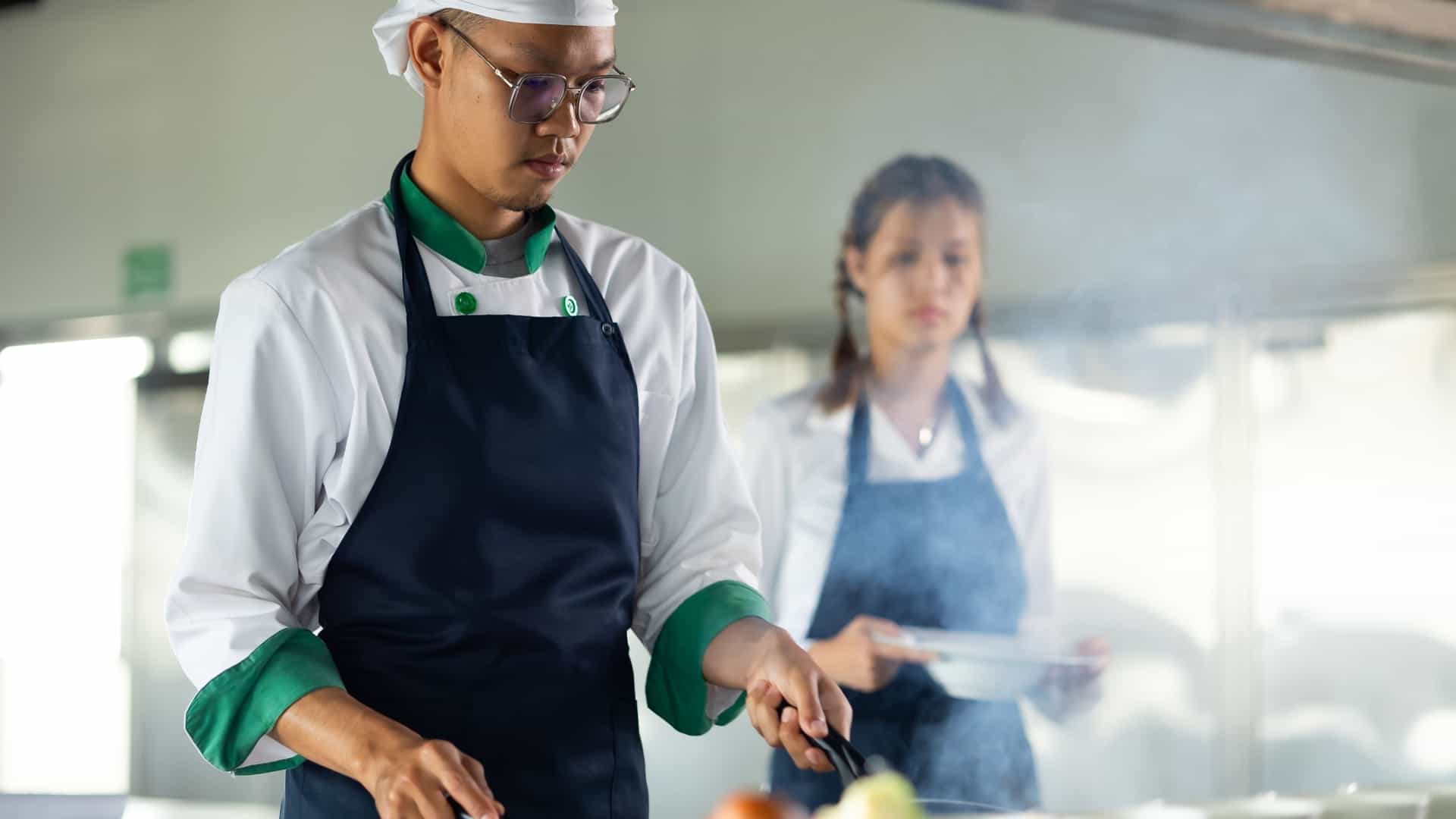  This is a close up portrait of an Asian Thai male professional chef cooking a meal in a culinary classroom. The image captures the chef during a cooking class or Chef lessons in school, concentrating on the ingredients and holding cooking utensils in the kitchen.